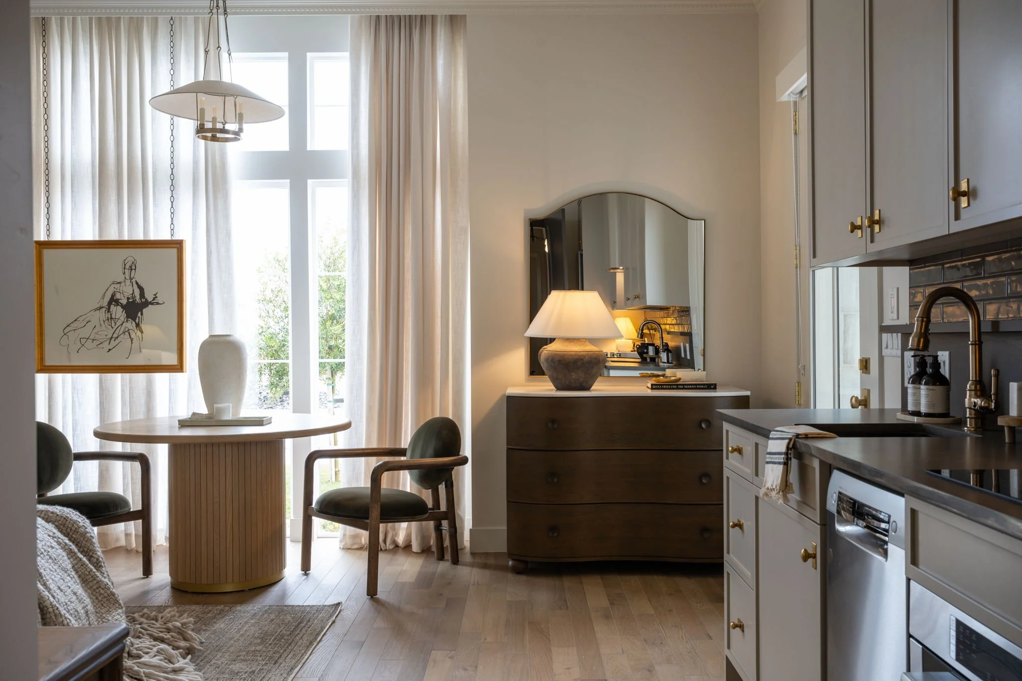 A cozy kitchen and dining area with a round white table, two green chairs, and a window with curtains letting in natural light; a sideboard with a table lamp and a mirror above it; kitchen cabinets with gold knobs, a sink with a gold faucet, dishwash