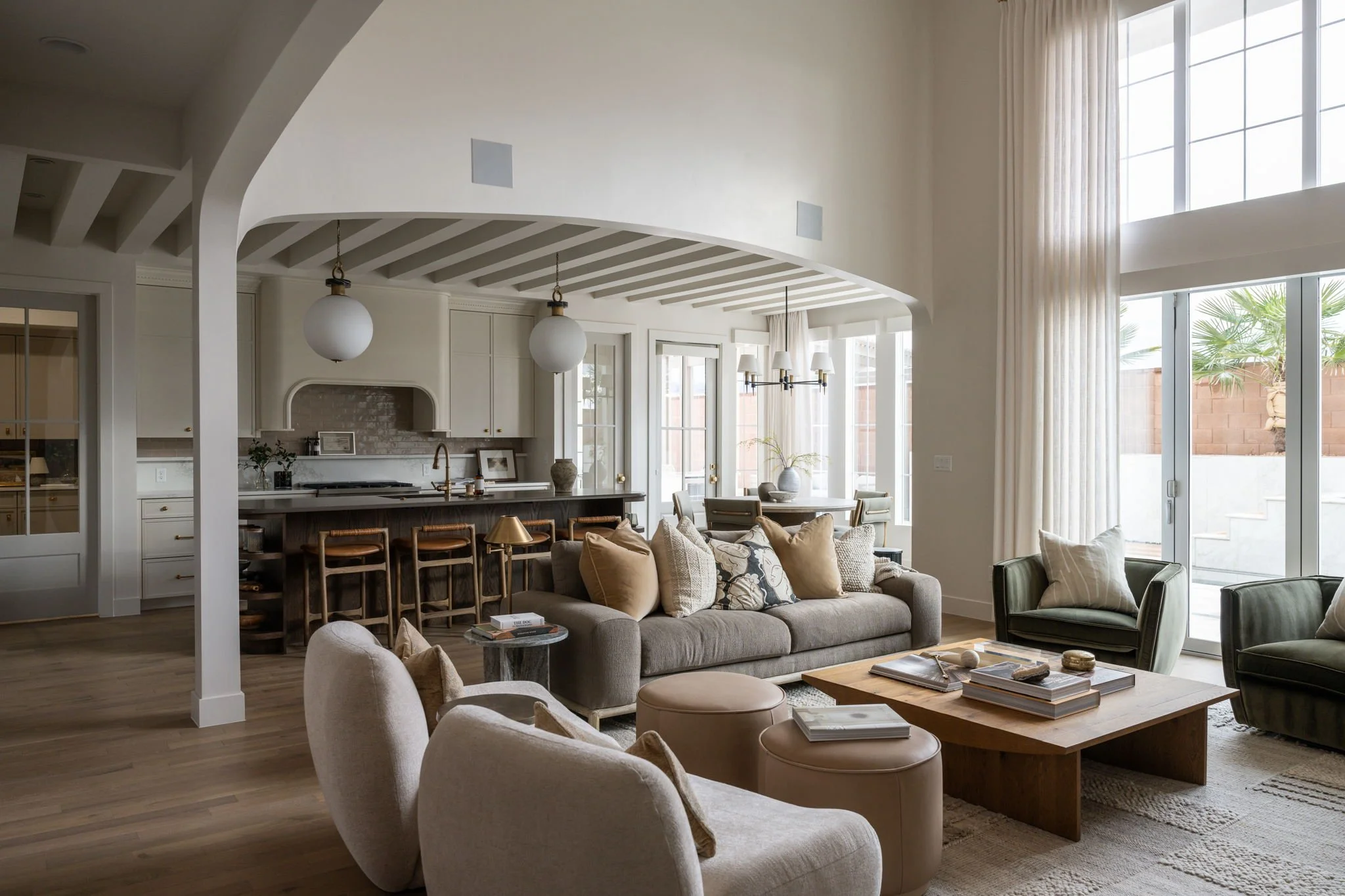 Bright living room with beige sofas, a wooden coffee table, and large windows with cream curtains, overlooking a small outdoor patio with plants.