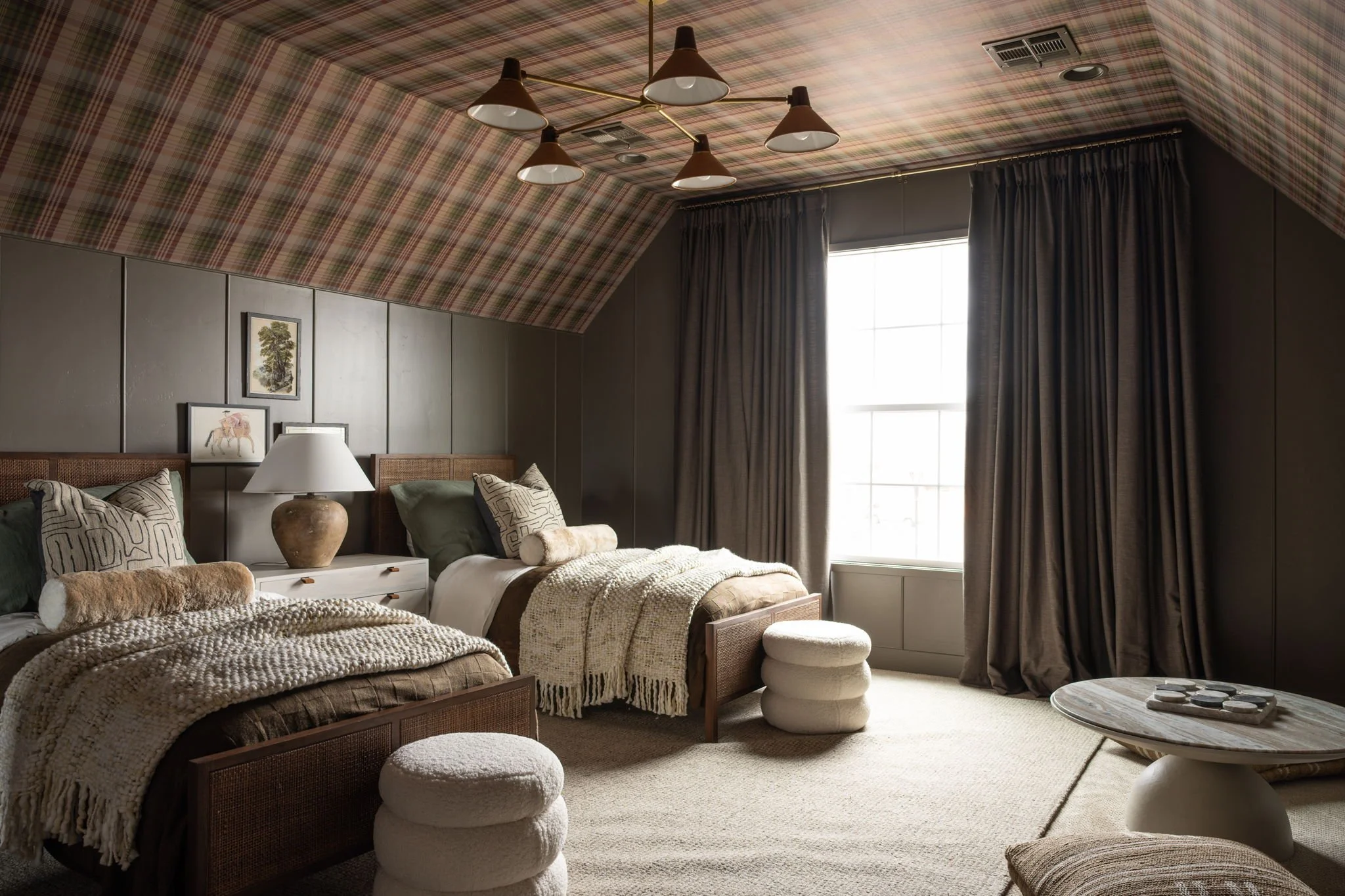 A bedroom with two beds, a bedside table with a lamp, framed pictures on the wall, a window with dark curtains, and a round table with decorative objects.