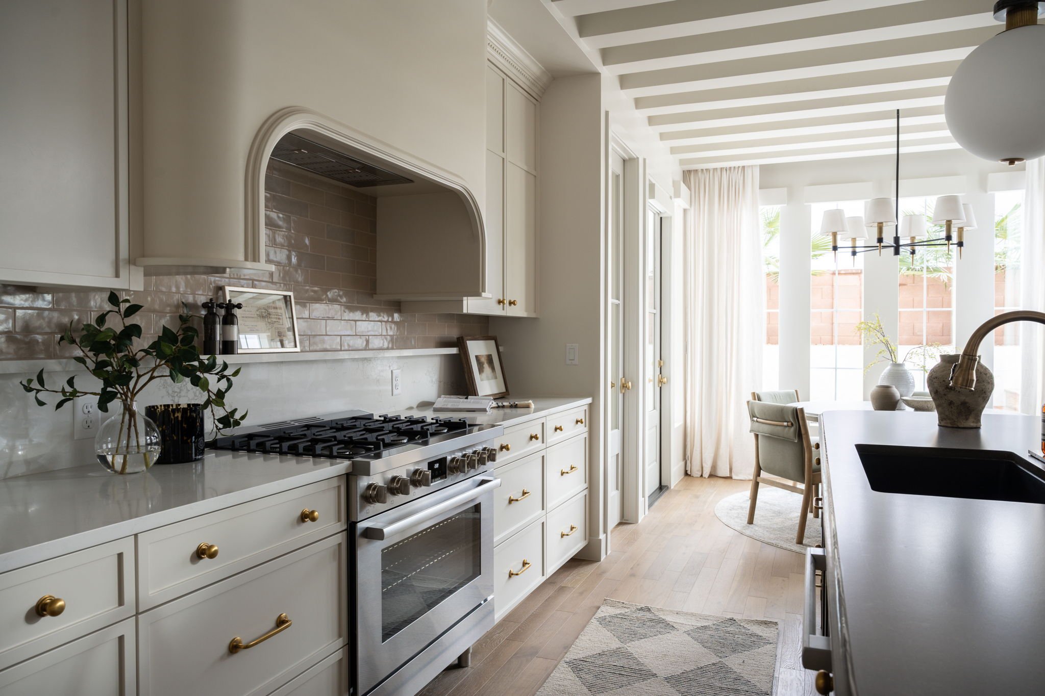 Bright kitchen with white cabinets, beige subway tile backsplash, and a gas stove. A vase with greenery and black containers sit on the countertop. Light wood floors and a chandelier over a dining area with chairs and a round rug are visible near lar