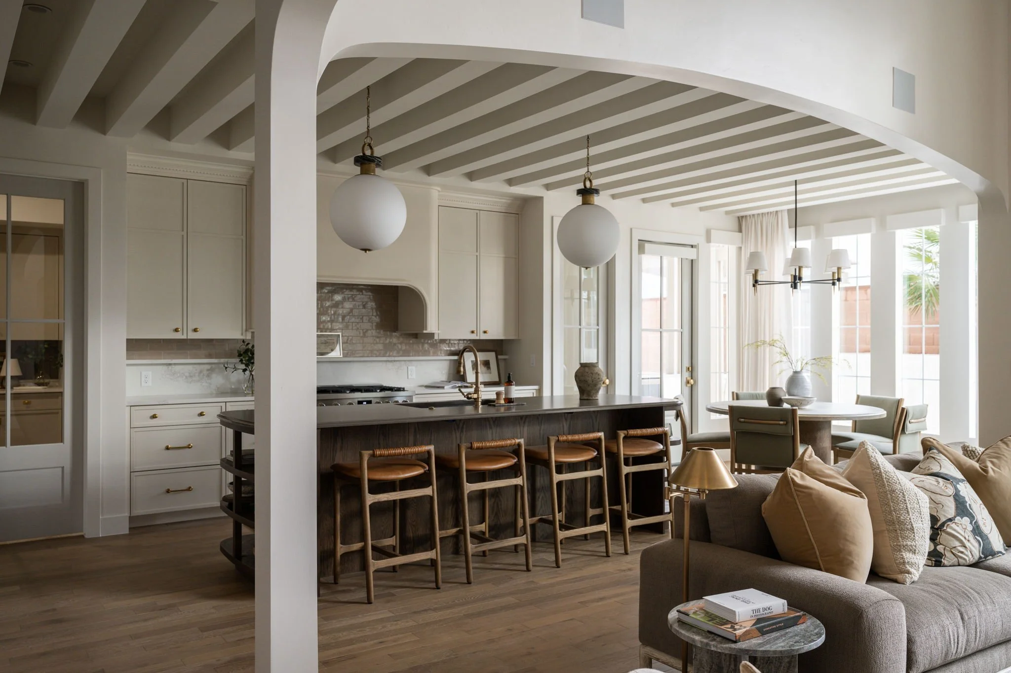 Open-concept kitchen and living room with white cabinets, wooden bar stools, beige sofa with pillows, round dining table with chairs, large windows, and modern light fixtures.