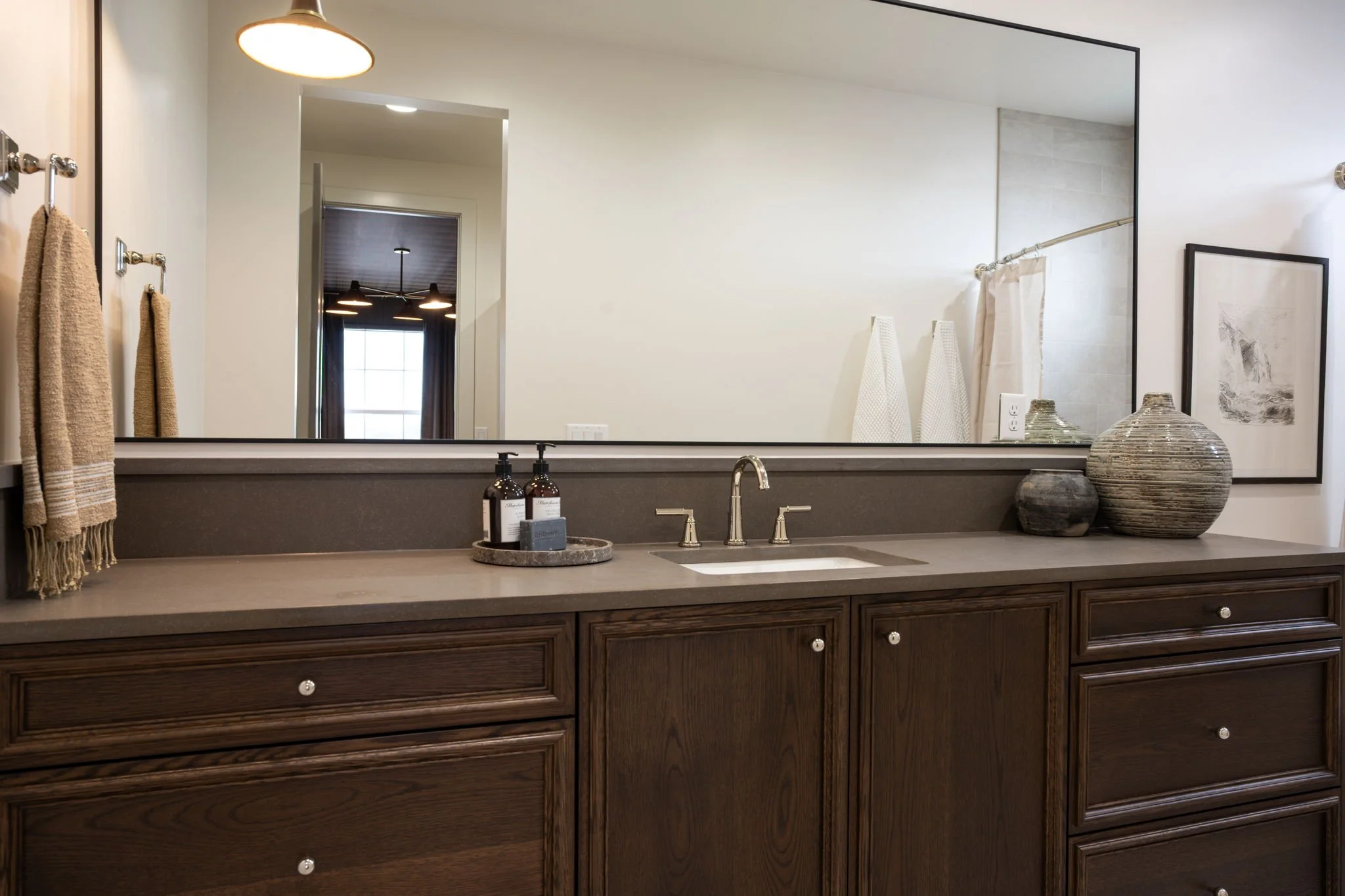 Bathroom vanity with a large mirror, dark wood cabinets, and decorative vases, with a view of the bathroom and bedroom through the door.