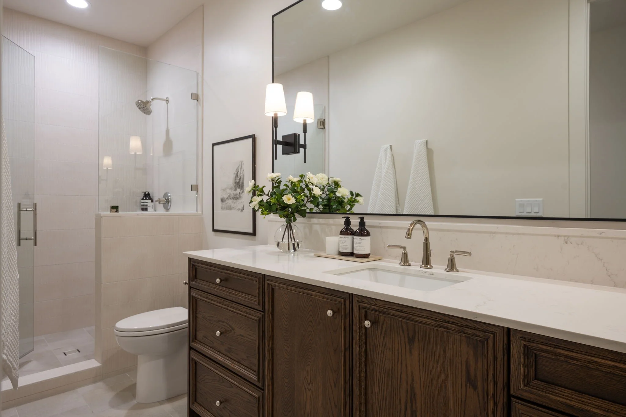 Modern bathroom with a large mirror, a wooden vanity with a marble countertop, a vase of white flowers, soap dispensers, a wall-mounted light fixture, towels hanging on the wall, a glass-enclosed shower with a showerhead, and a white toilet.