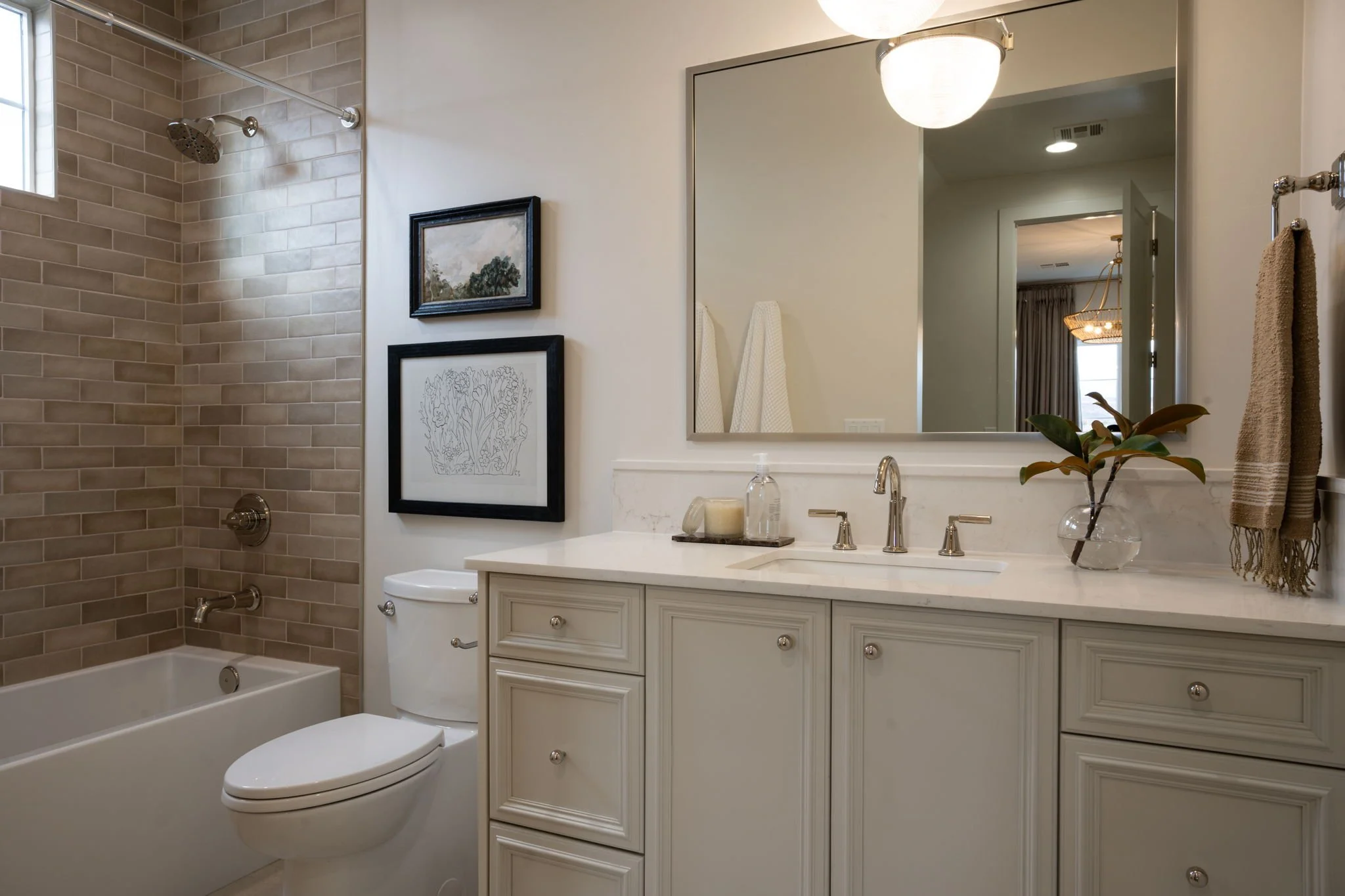 A bathroom with a bathtub and shower, a toilet, a large mirror above a white vanity with cabinets, a small plant in a glass vase, and framed artwork on the wall.