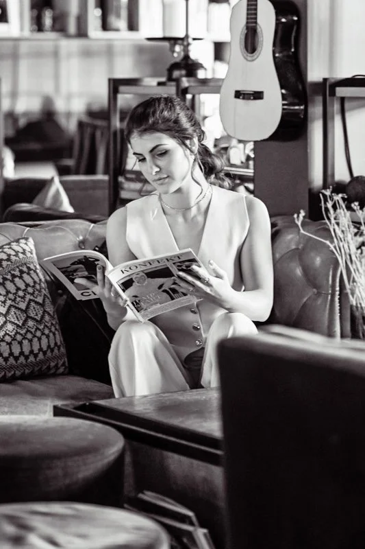 Une jeune femme assise sur un canapé en cuir, lisant un magazine dans un intérieur avec une guitare accrochée au mur derrière elle.