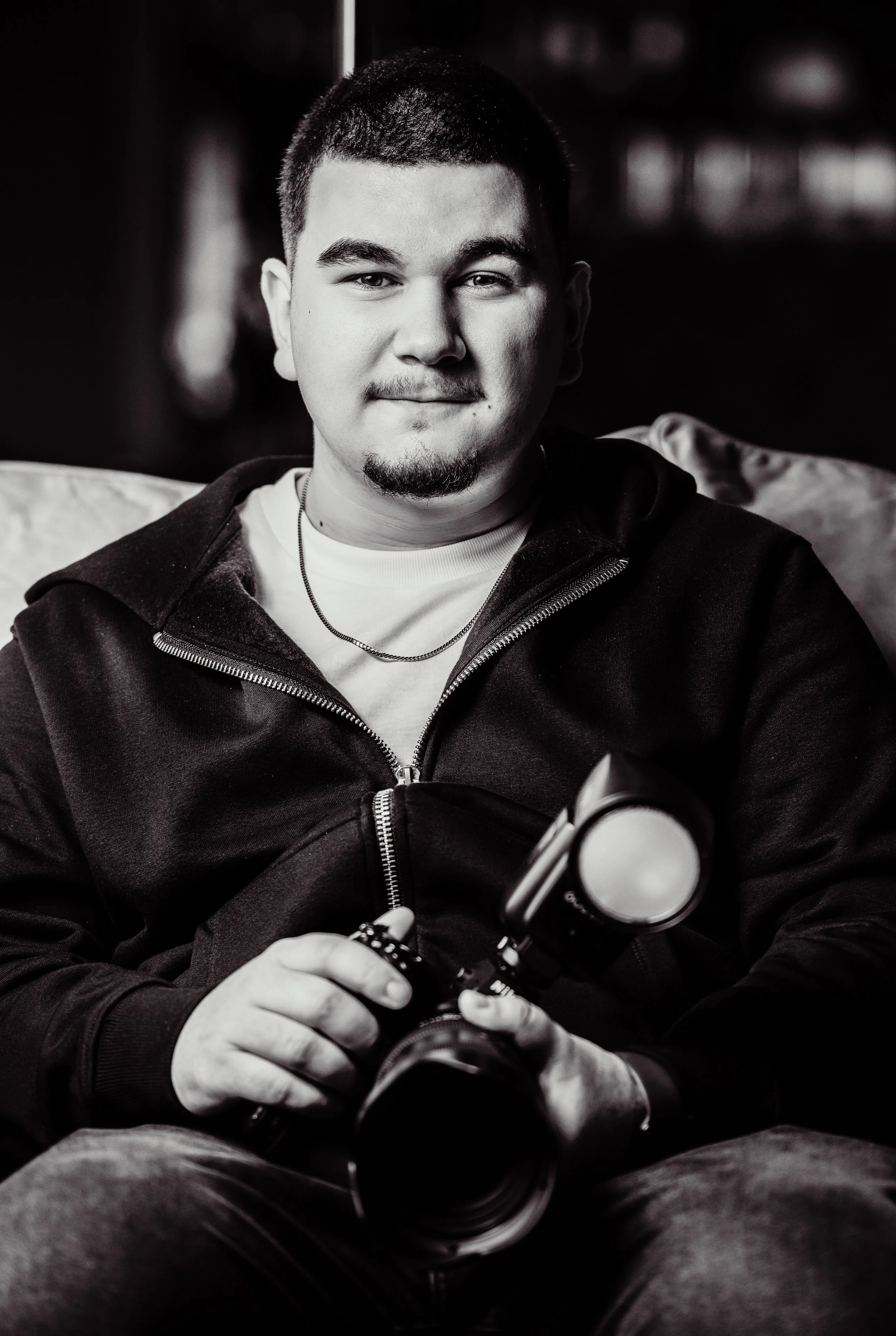 Un jeune homme souriant, tenant un appareil photo avec flash, assis sur un canapé, en noir et blanc.