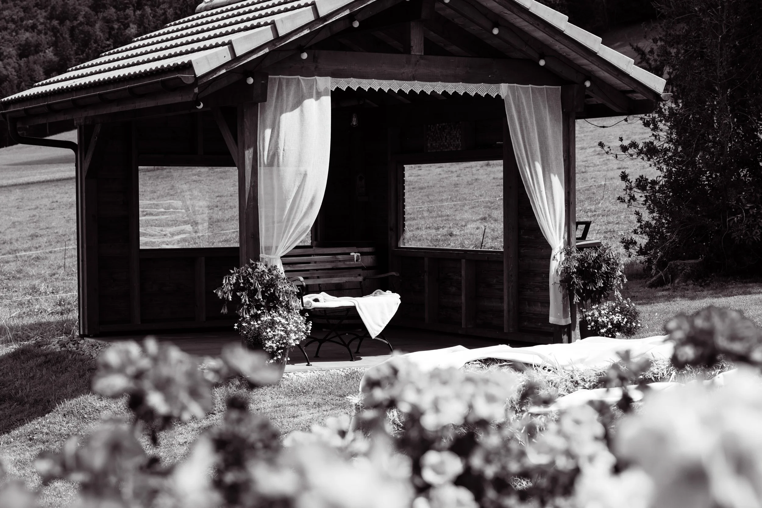 Une petite cabane en bois avec des rideaux blancs, située dans un champ avec des fleurs en premier plan.
