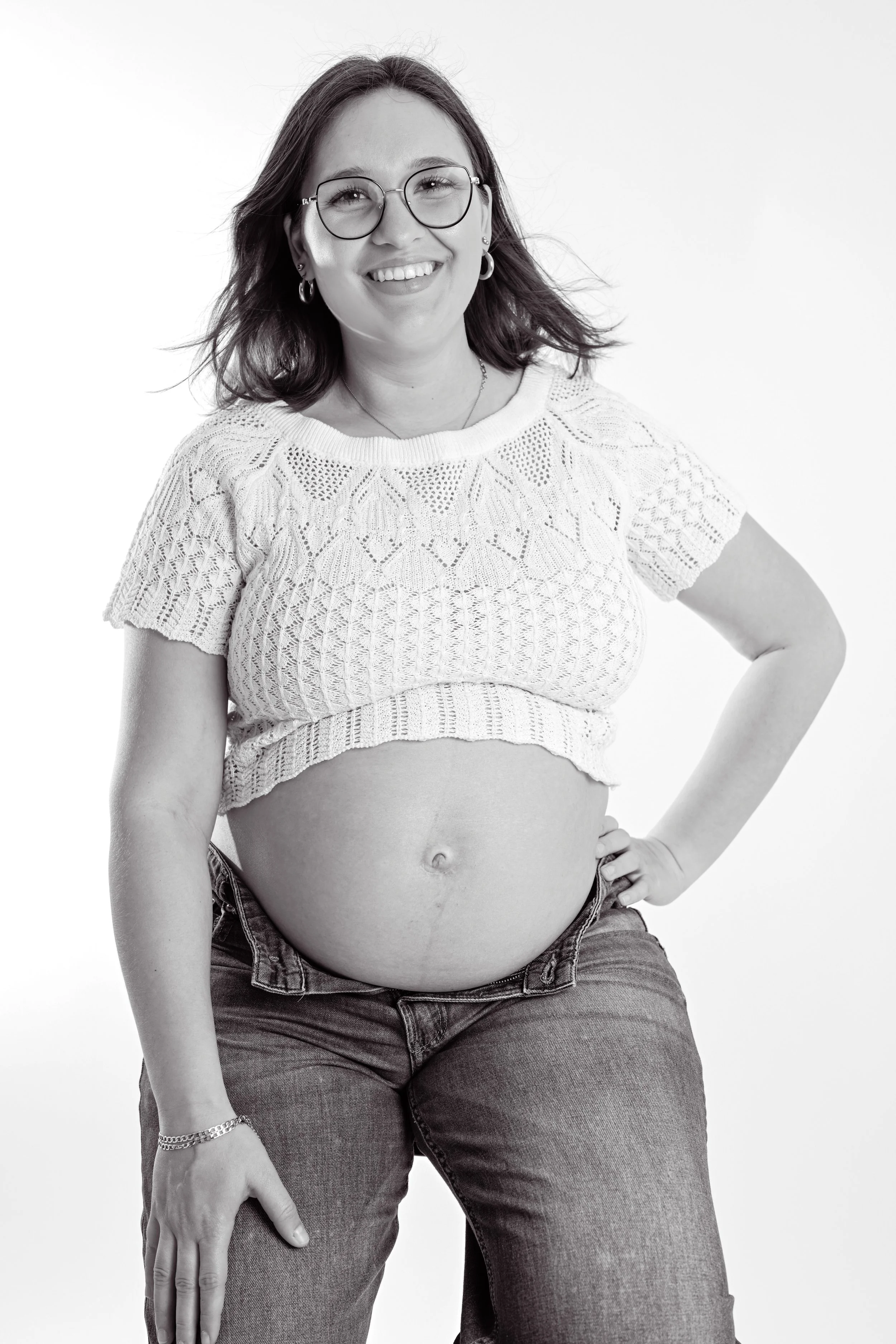 Une femme enceinte souriante portant des lunettes, un pull en crochet court et un jean, posant avec une main sur la taille, dans un fond blanc.