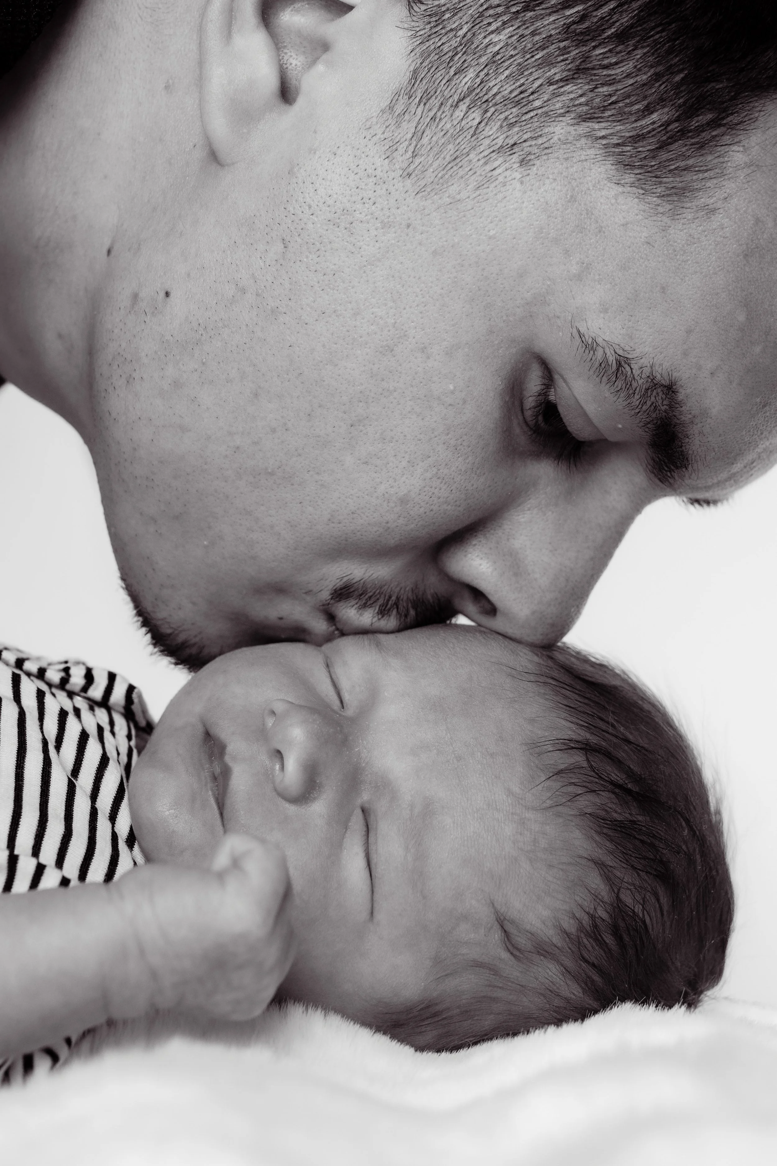 Un homme jeune embrassant un bébé endormi sur le front, en noir et blanc.