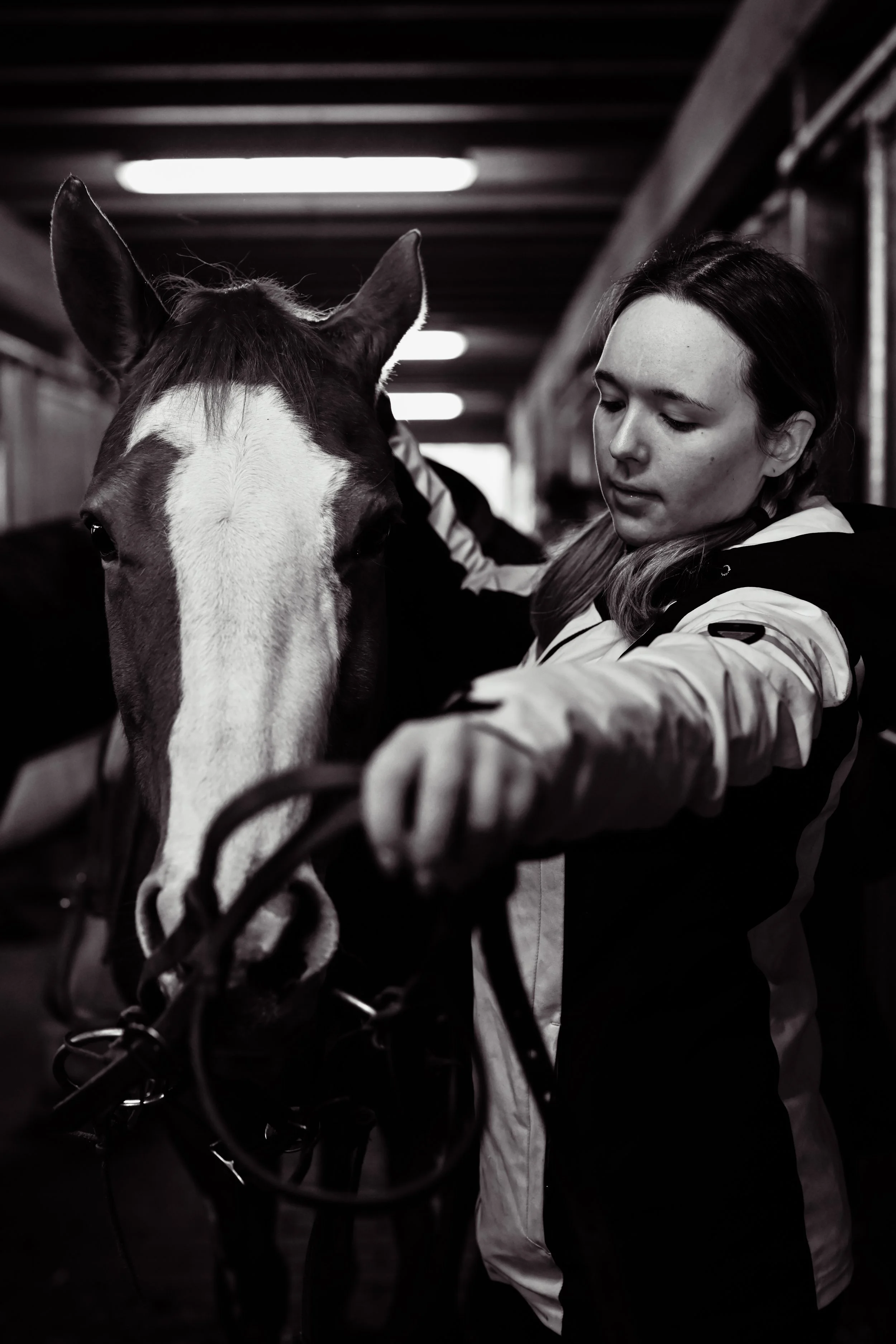Une jeune femme s'apprête à monter un cheval dans une écurie, en noir et blanc.