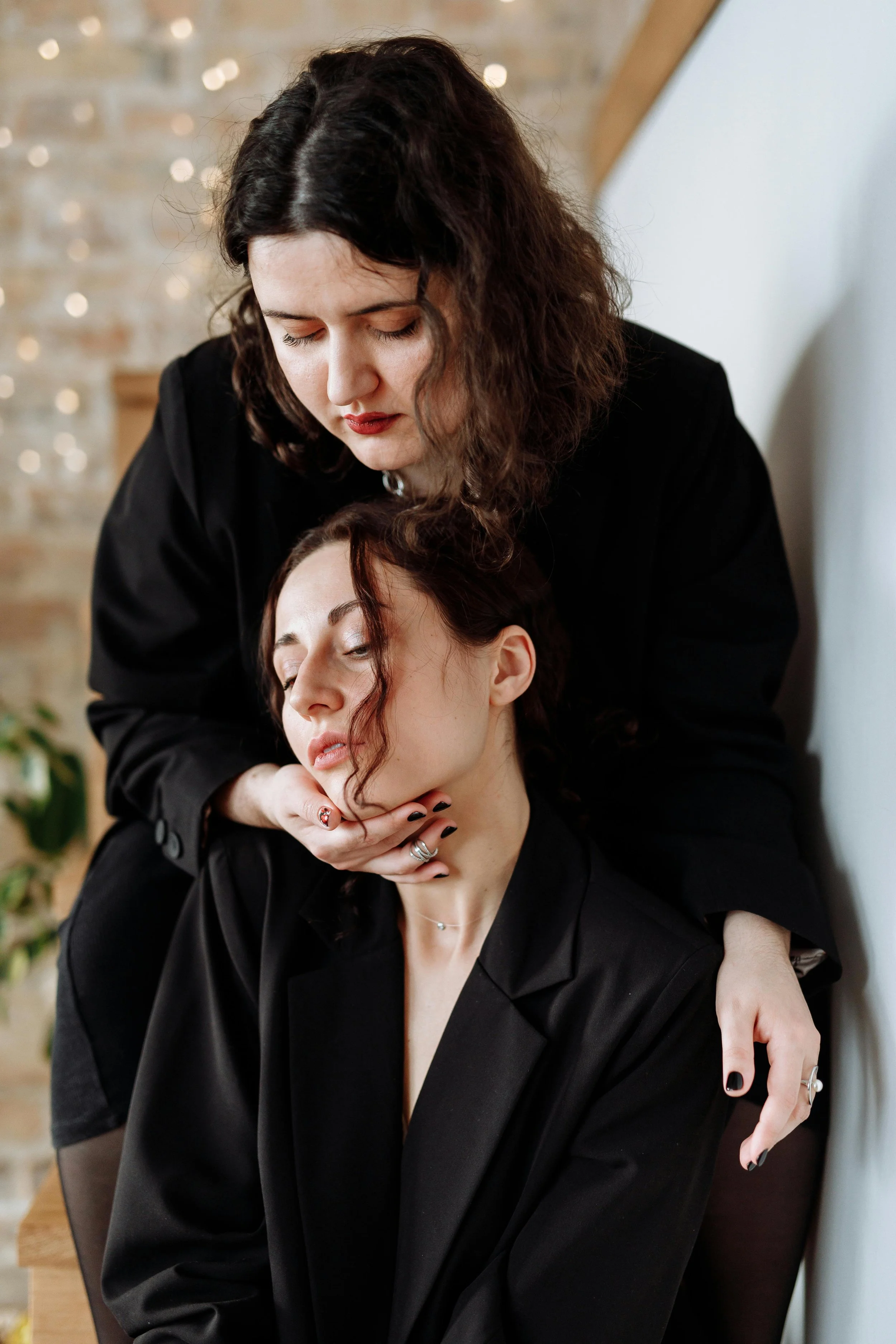 A makeup artist applies makeup to a woman with closed eyes, who is sitting with her head tilted back, against a background with string lights and a brick wall.