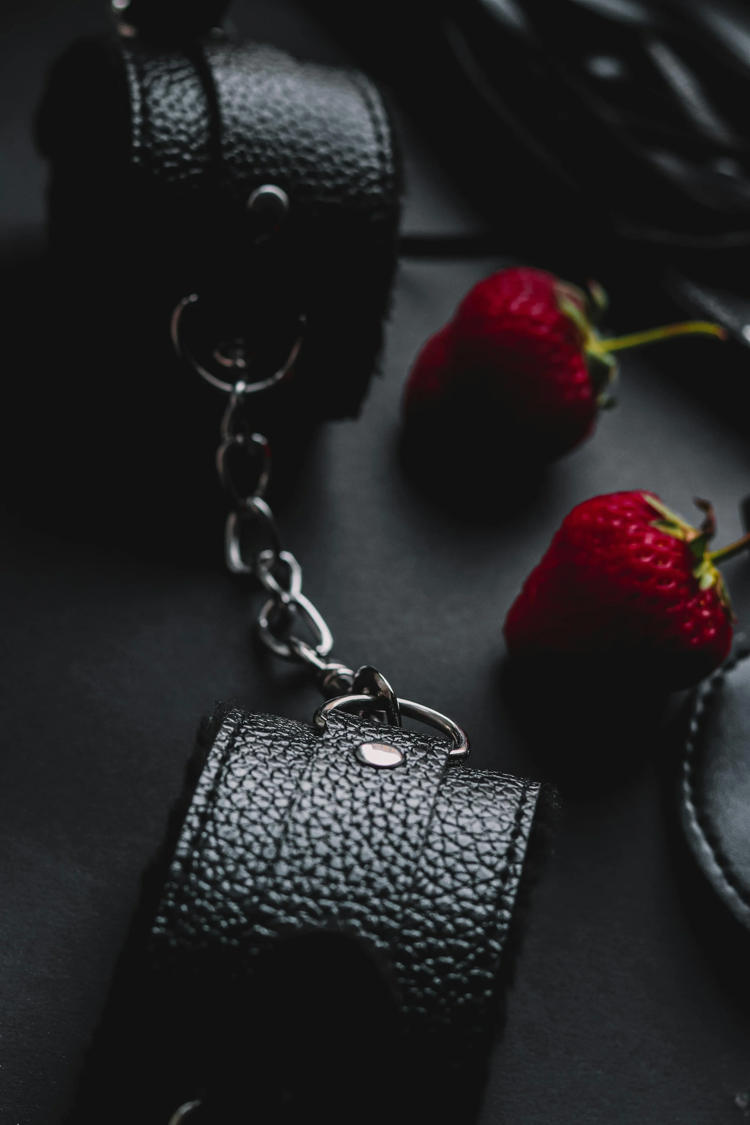 Close-up of black leather accessories with silver chain and two strawberries on a dark surface.
