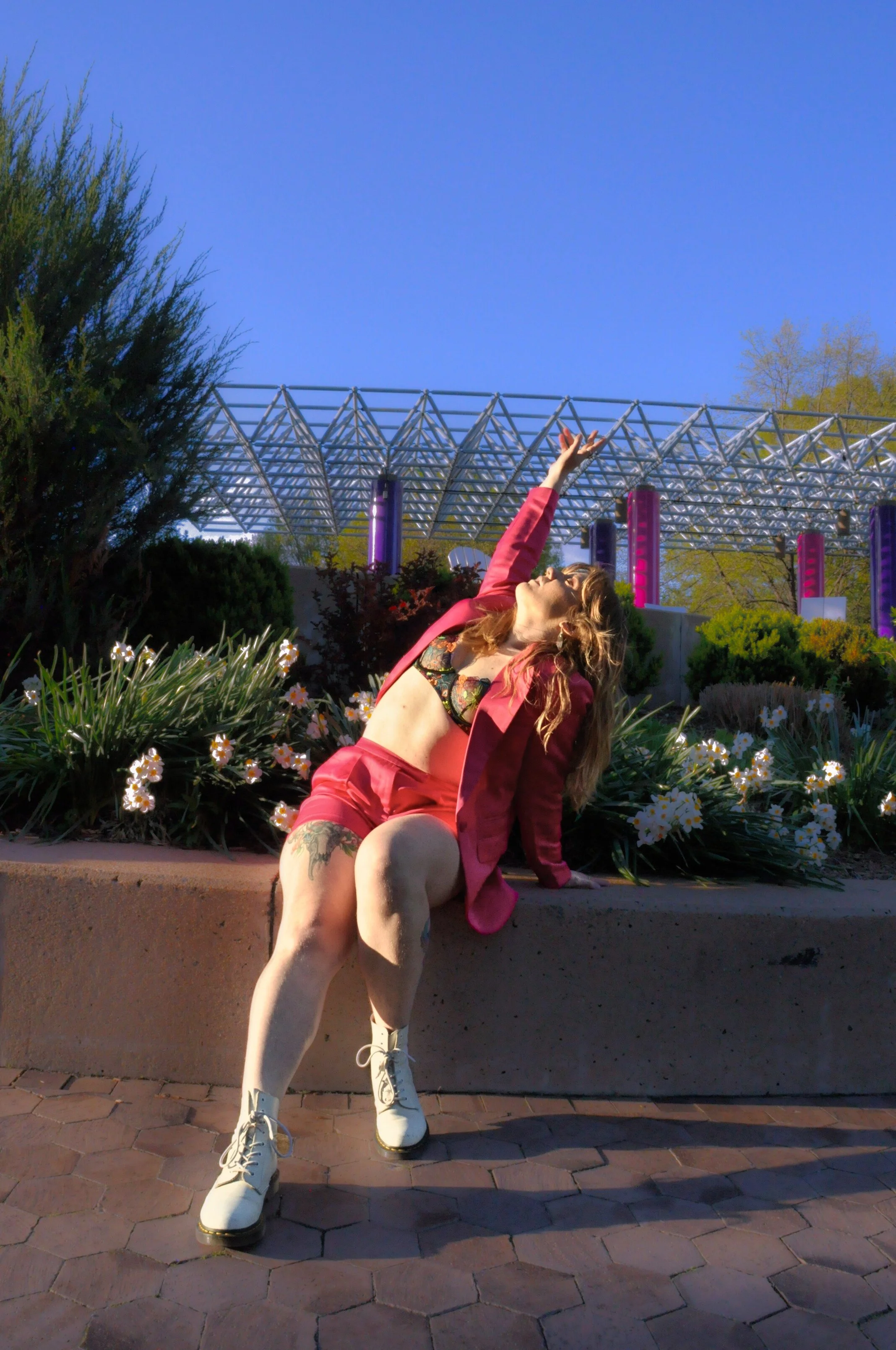 A woman sitting on a planter with flowers, wearing a pink blazer, lingerie top, shorts, and white boots, striking a pose with her right arm extended upward against a clear blue sky and modern bridge structure in the background.