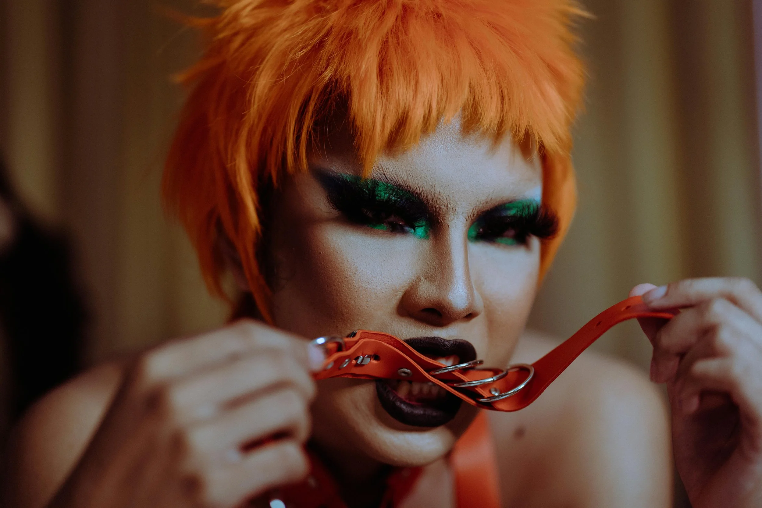 Person with vivid orange hair, heavy makeup, green eyeshadow, and dark lipstick, biting an orange pair of glasses.