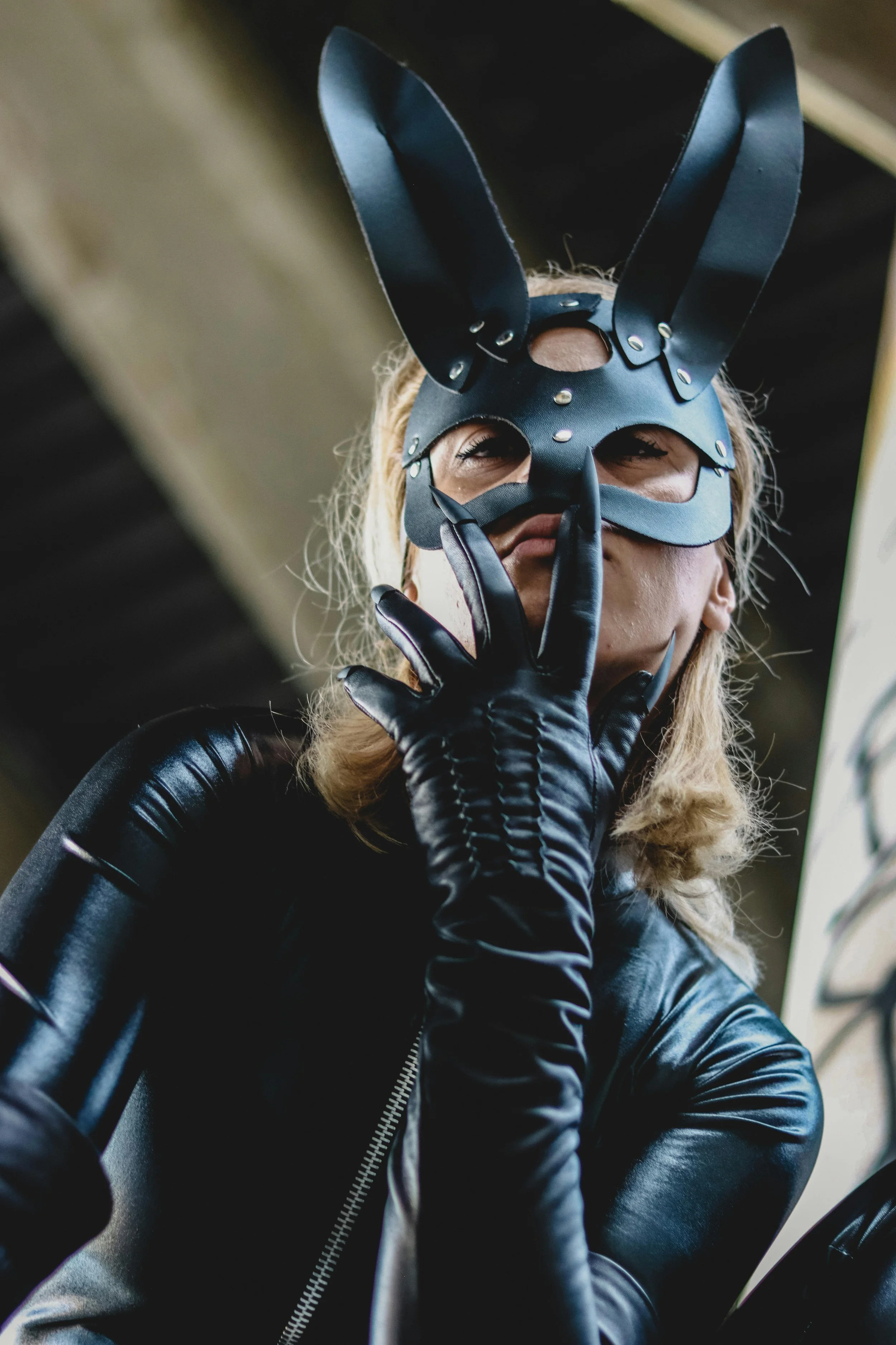 A person wearing a black bunny mask with tall ears, black gloves, and a black leather jacket making a shushing gesture.