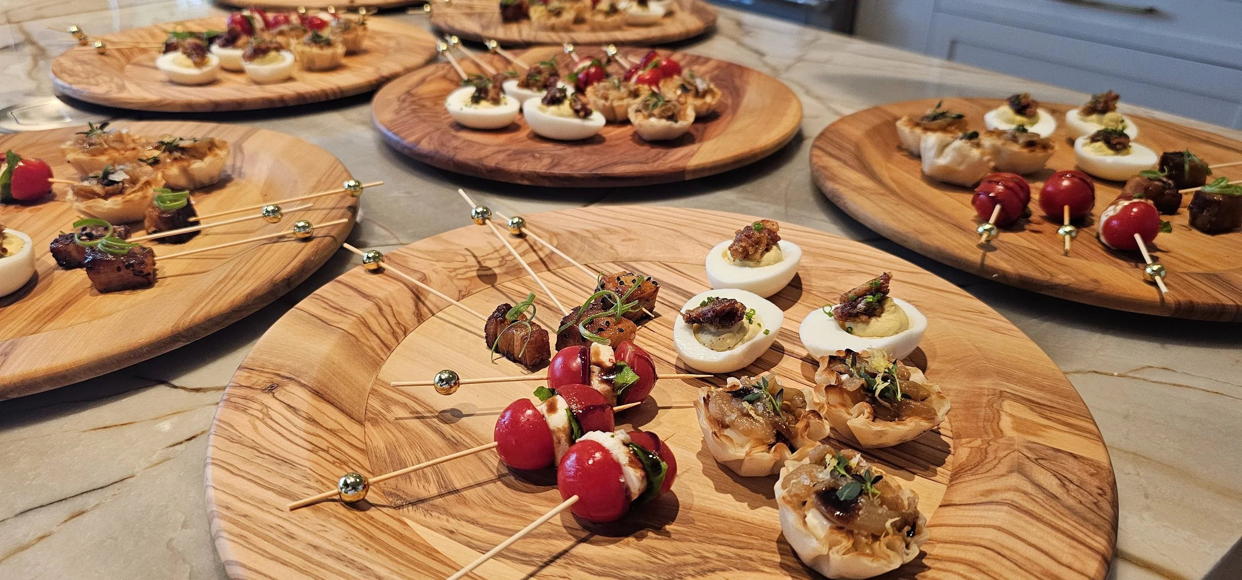 Assorted appetizers served on wooden plates, including deviled eggs topped with bacon and herbs, cherry tomatoes with diced cheese and herbs on skewers, and small pastry cups filled with savory toppings, all arranged on a marble countertop.