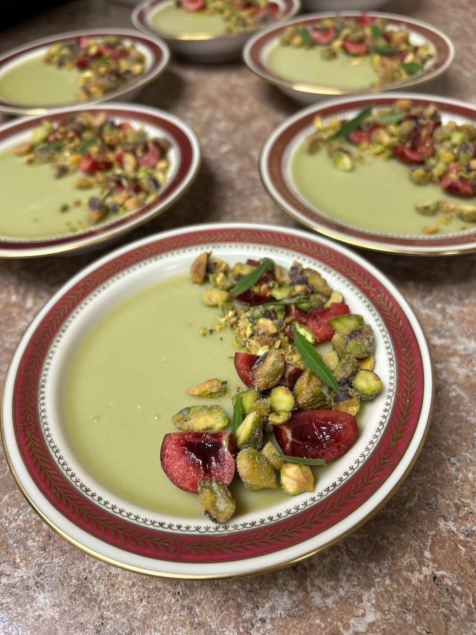 Several plates of green-colored dessert garnished with chopped pistachios, red fruit, and green herbs on a marble surface.