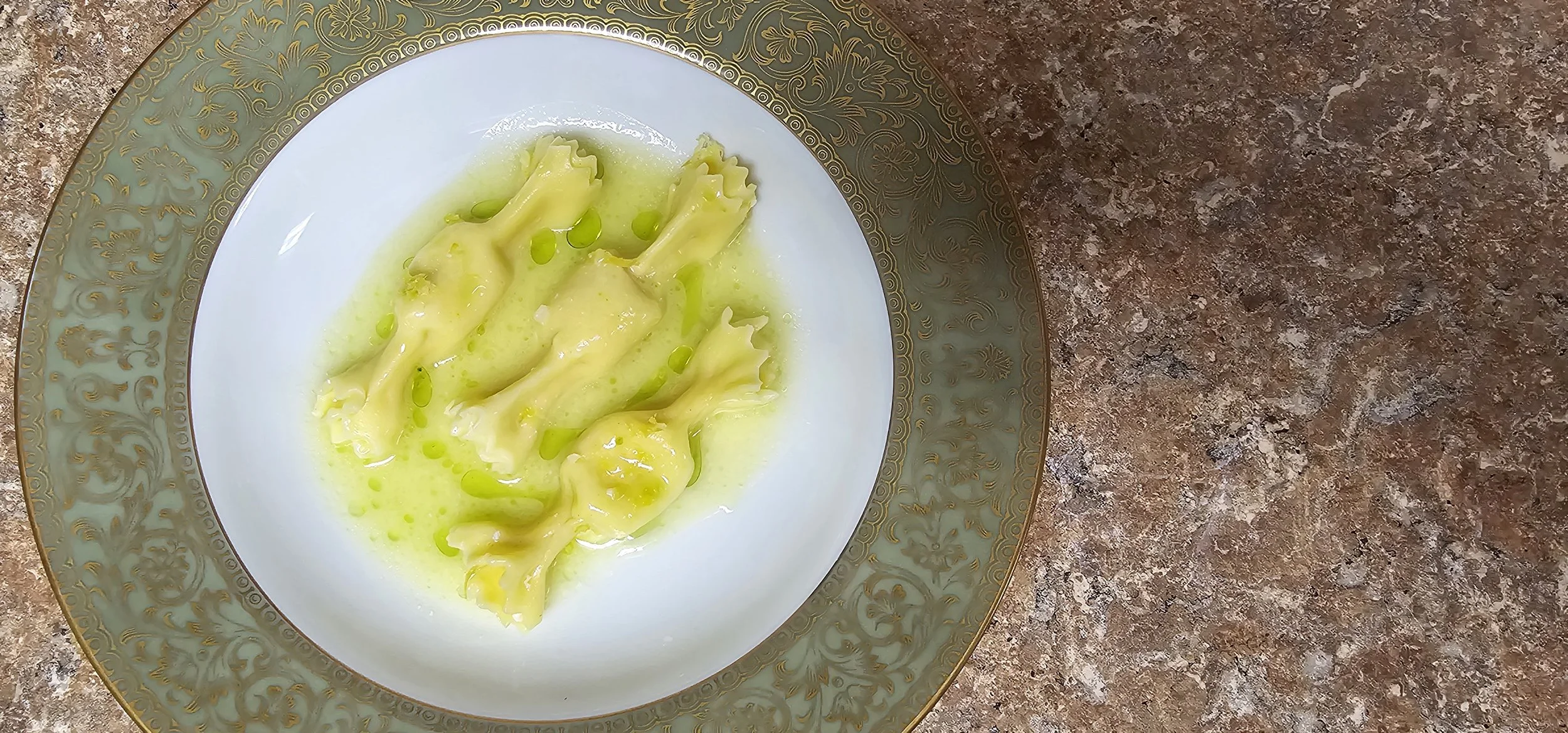 Plate with three ravioli pasta pieces in a green sauce, garnished with small green droplets, on a decorative gold and white plate on a brown marble countertop.