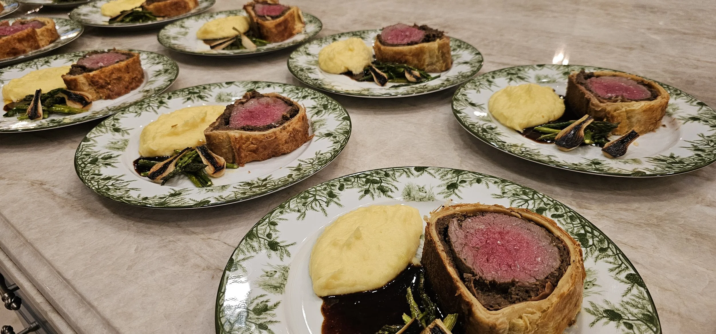 Multiple plates with sliced beef Wellington, mashed potatoes, grilled vegetables, and dark gravy.