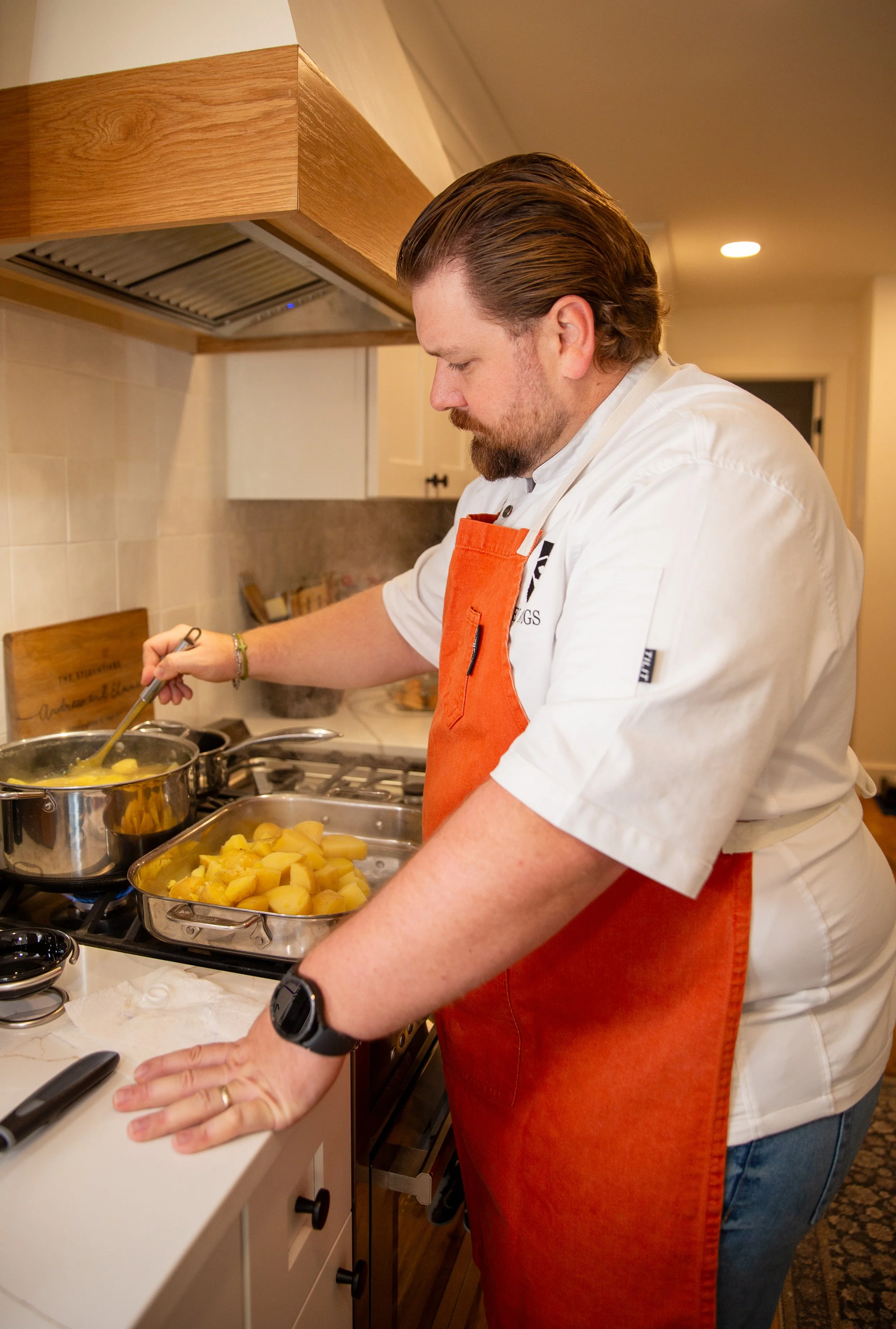 A man wearing a white chef's coat and an orange apron is cooking on a stove, stirring a pot of yellow potatoes or similar vegetables in a kitchen.