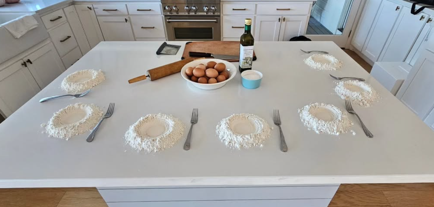 Kitchen island with six sets of flattened dough circles surrounded by flour, with utensils, a bowl of eggs, a rolling pin, a bottle of oil, and a small container of flour on the counter.