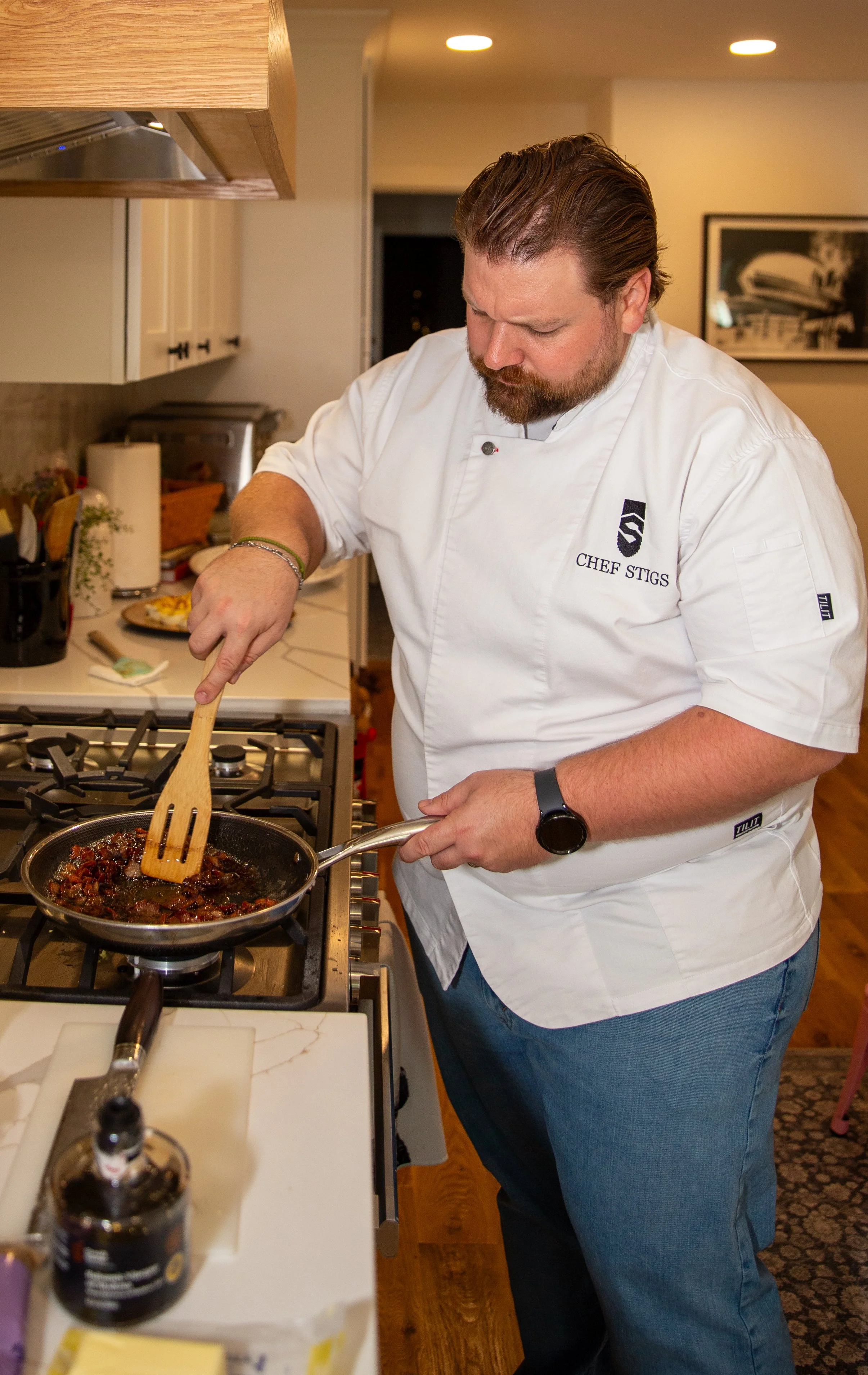 A chef wearing a white coat with the name "Chef Stigs" is cooking on a stovetop, stirring food in a frying pan with a wooden spatula.