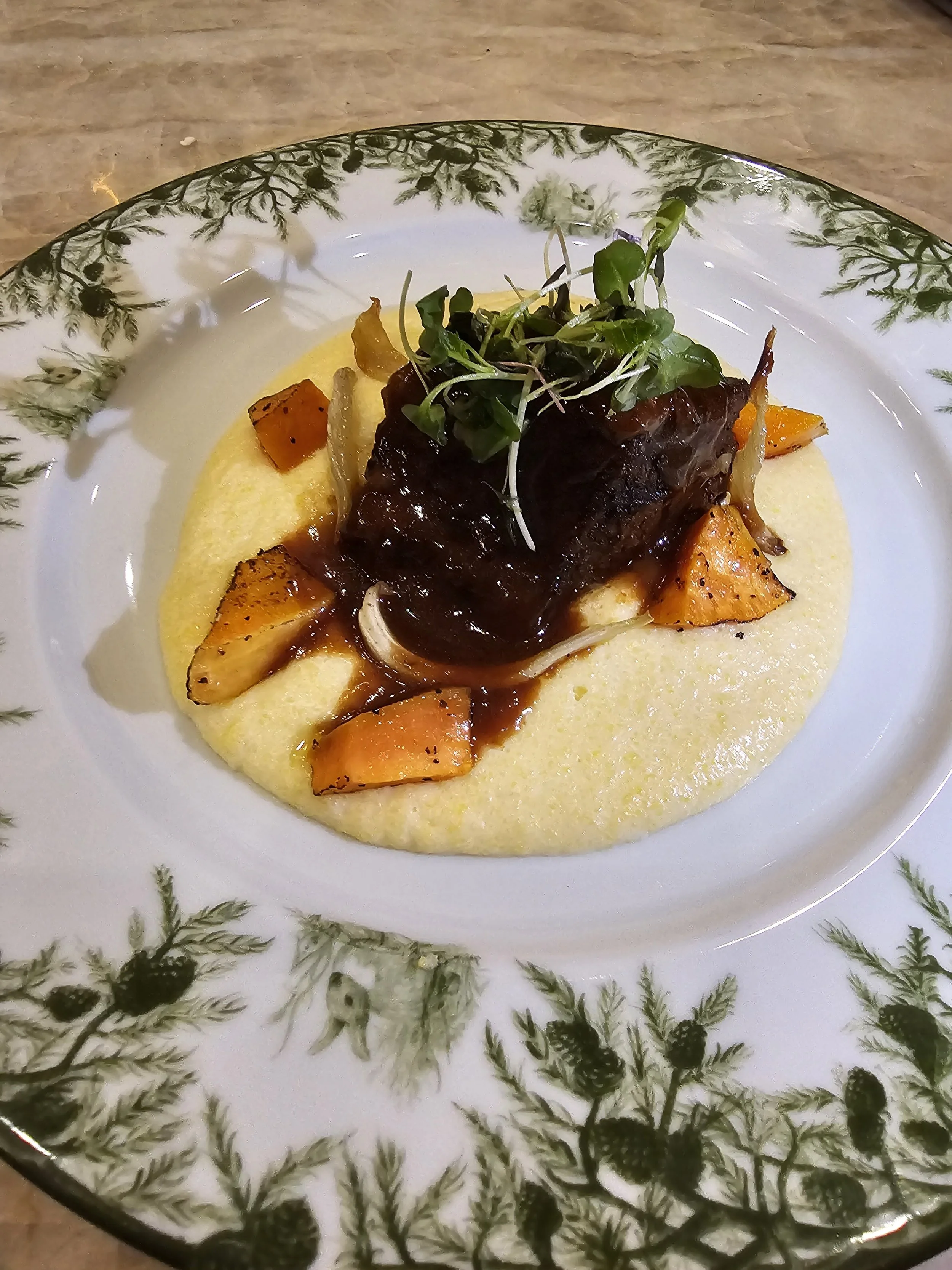 Plate of food with mashed potatoes topped with roasted carrots, a braised beef short rib, gravy, and microgreens on a decorative plate.
