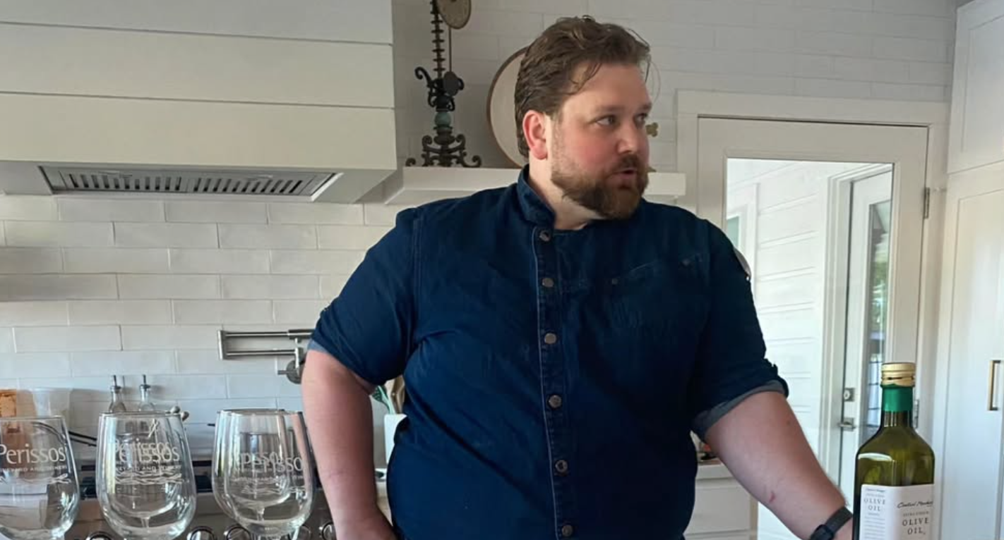 Man in a blue shirt standing in a kitchen with wine glasses and a bottle of olive oil on the counter.