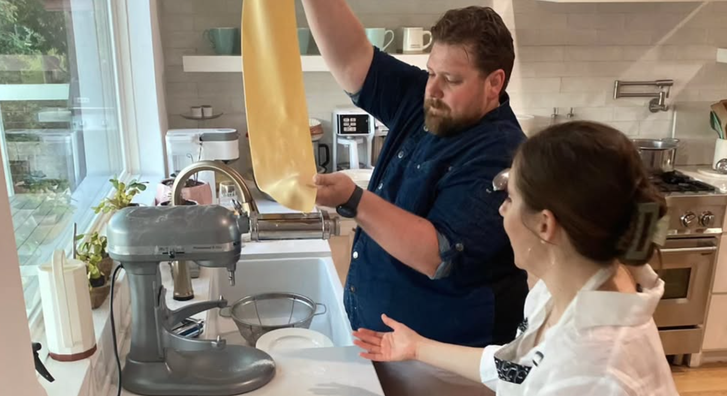 Man and woman making pasta in a kitchen, man pouring pasta dough into a machine, woman gesturing toward it.
