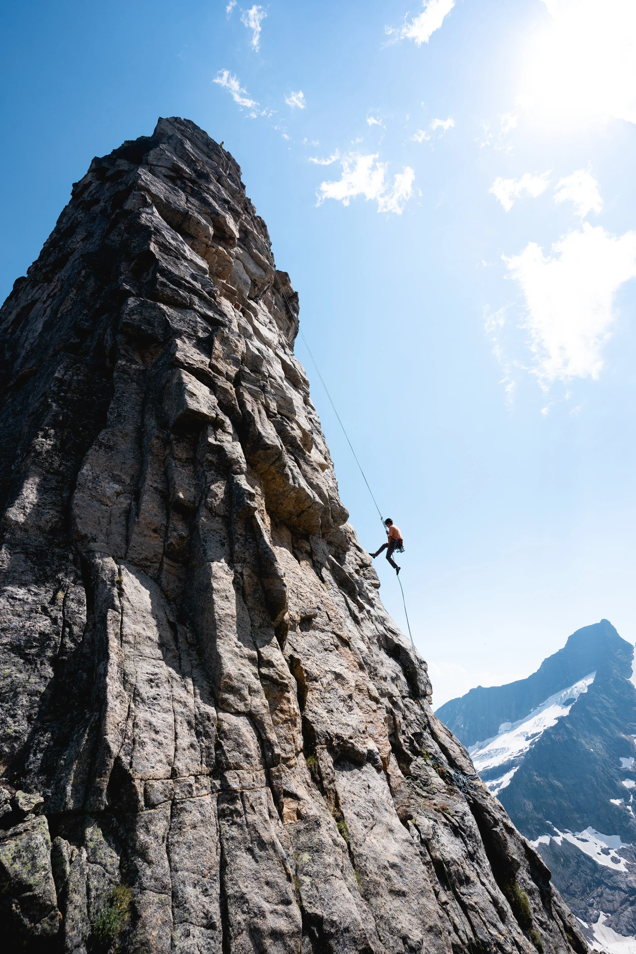 A climber hangs from a rope on a steep, rocky mountain slope with snow-capped peaks in the background under a partly cloudy sky.