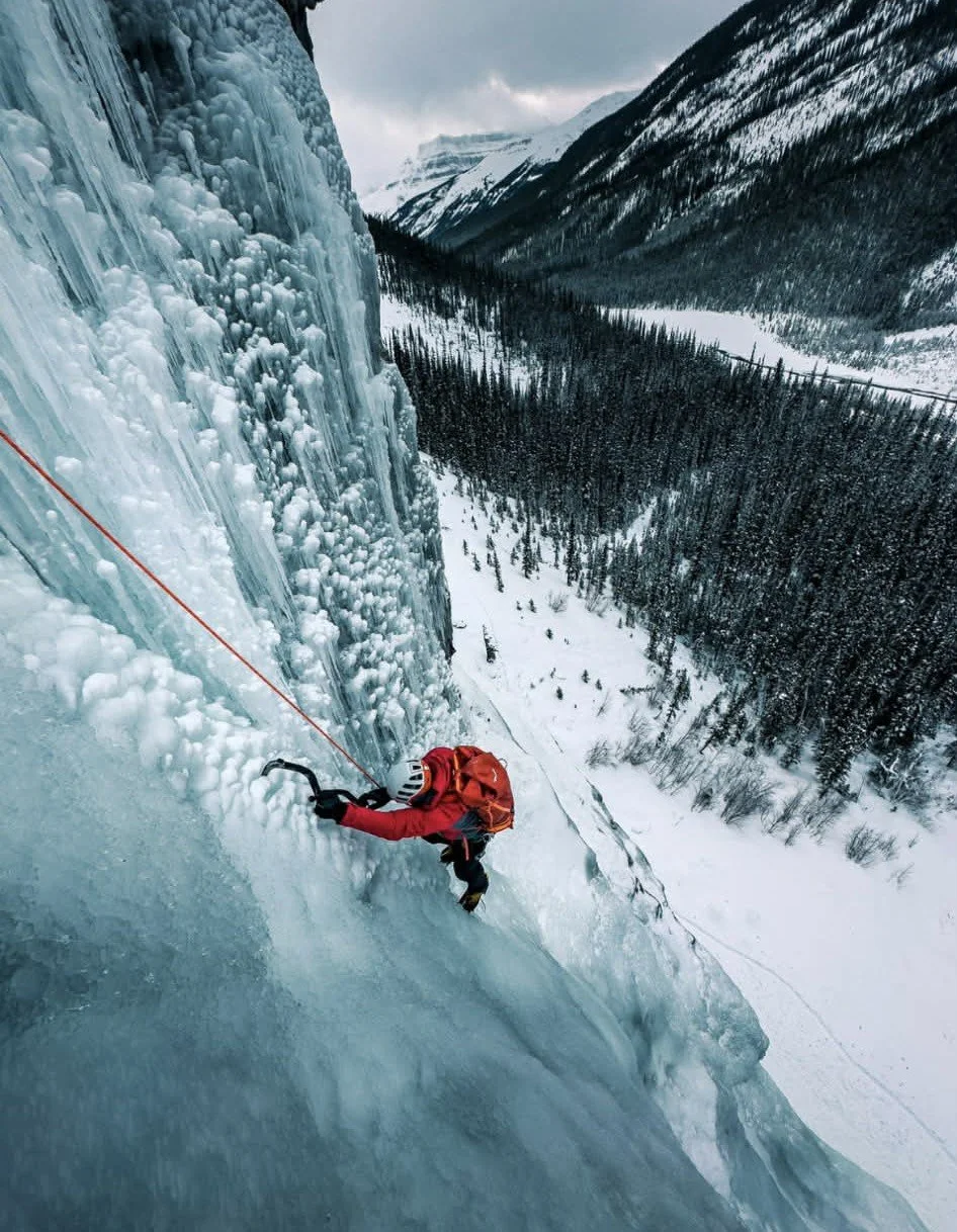 A person wearing a red jacket and helmet is ice climbing on a large ice formation in a snowy mountain landscape with a valley and forest in the background.