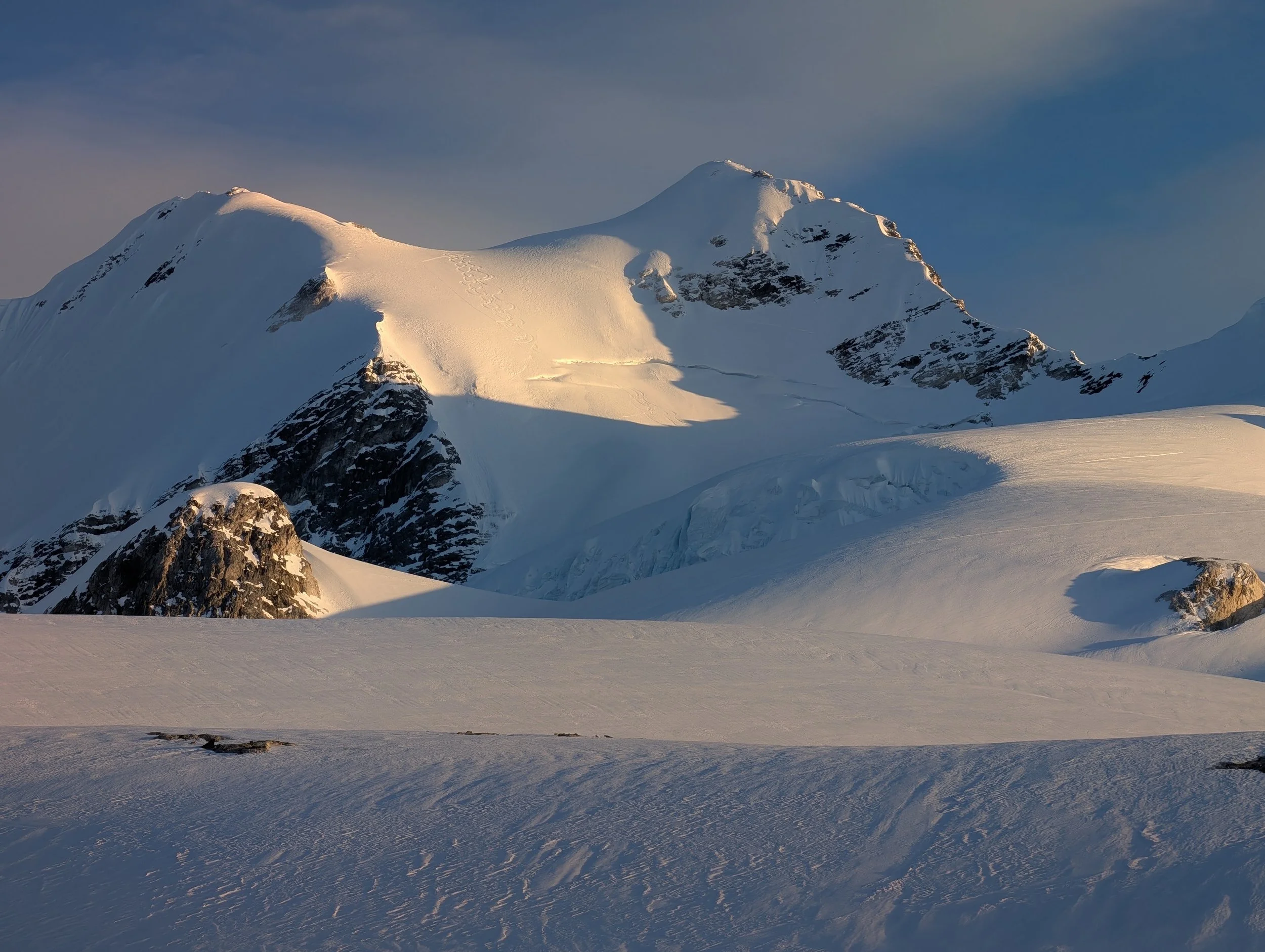 Snow-covered mountain peaks with tracks in the snow and a vast snowfield in the foreground under a cloudy sky.