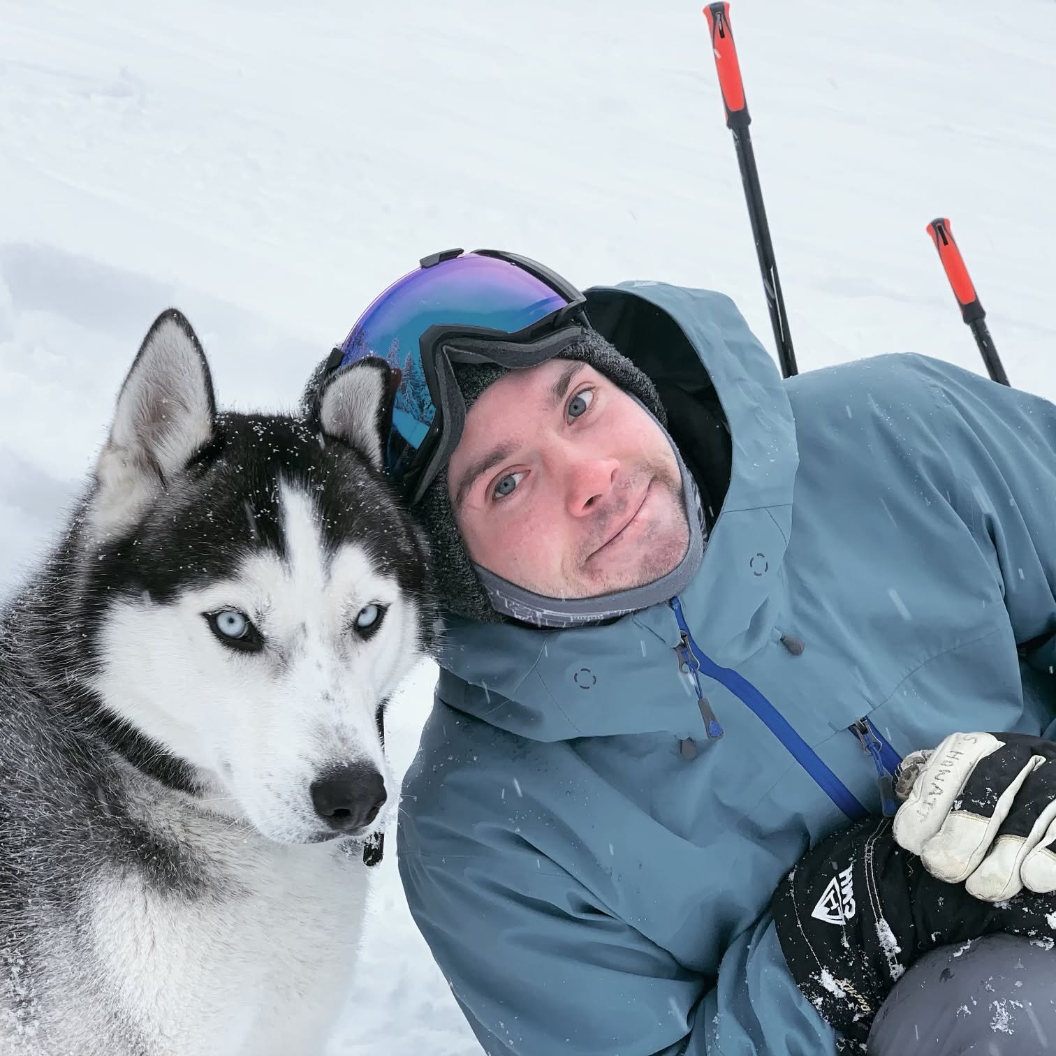 A man lying in the snow next to a Siberian Husky with blue eyes, both bundled in winter gear with snow on their faces during a snowy outdoor winter activity.