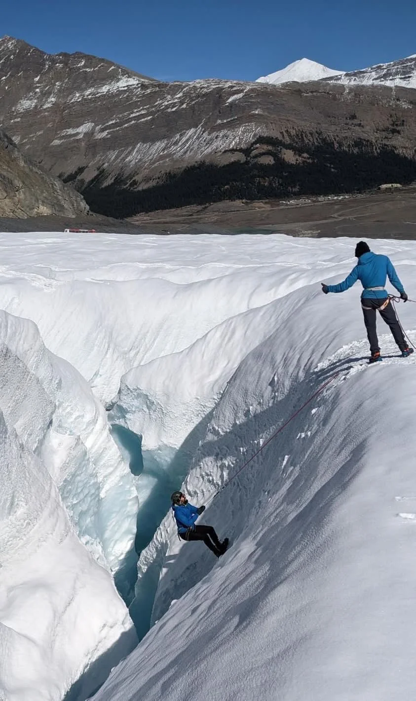 A person in a blue jacket and black pants is rappelling down a steep ice crevasse in a glacier, while another person in a similar outfit is standing on the ice holding the rope, in a mountainous, snow-covered landscape with mountains in the background.