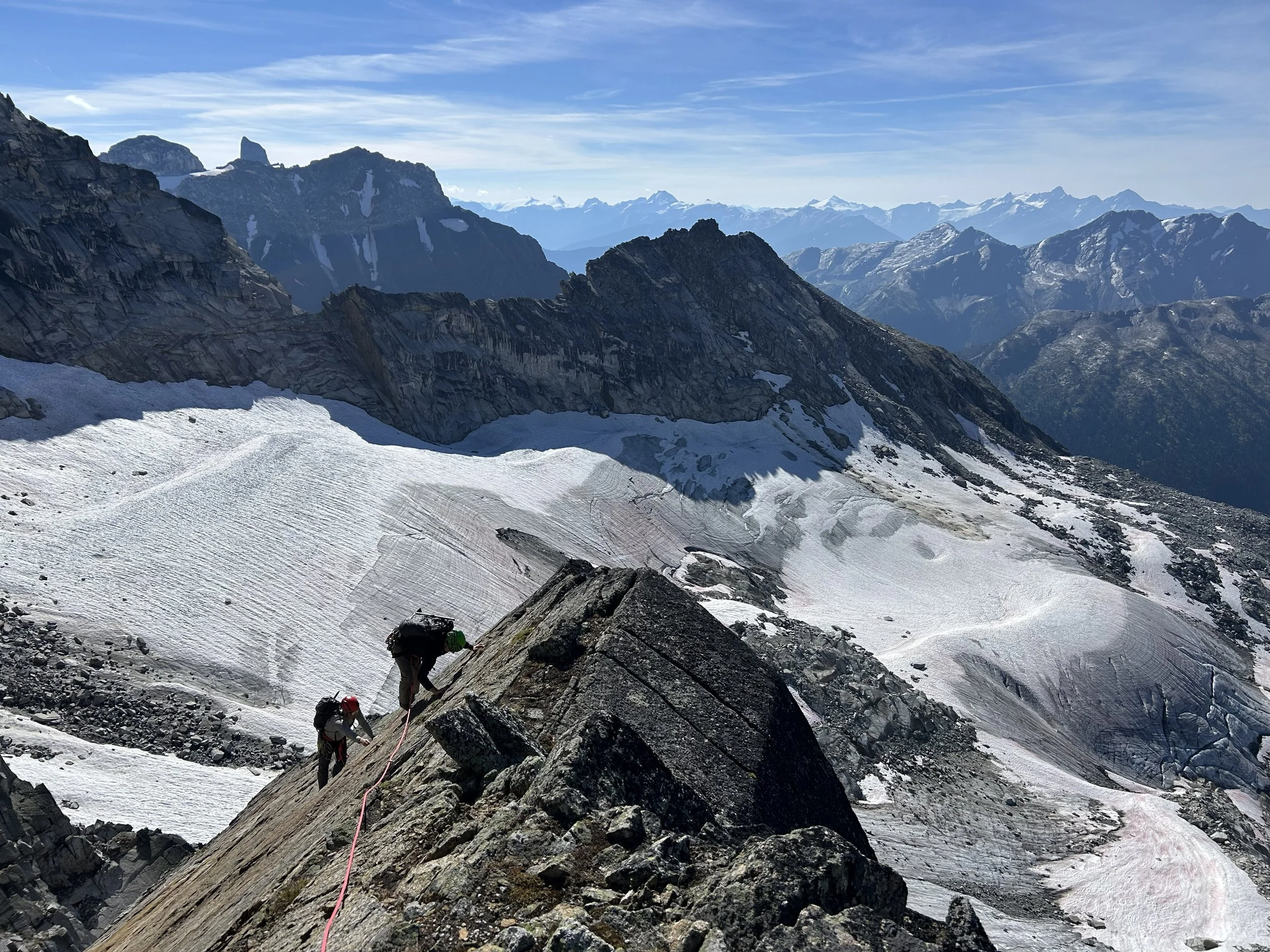 Two climbers in helmets and harnesses ascending a rocky mountain slope with snow patches, set against a backdrop of rugged mountain peaks and a clear blue sky.