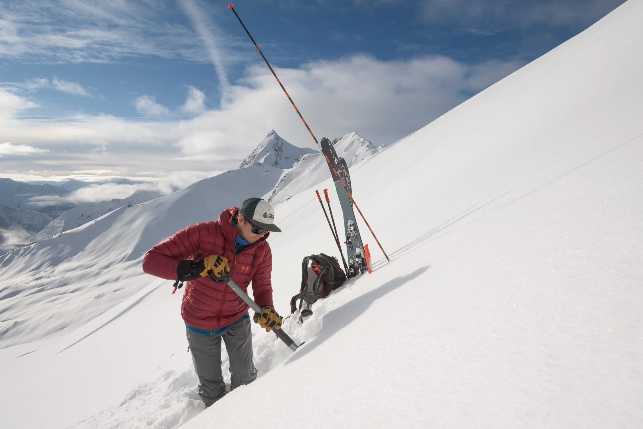 A person dressed in a red jacket and gray pants preparing ski equipment on a snowy mountain slope with a mountain range and cloudy sky in the background.