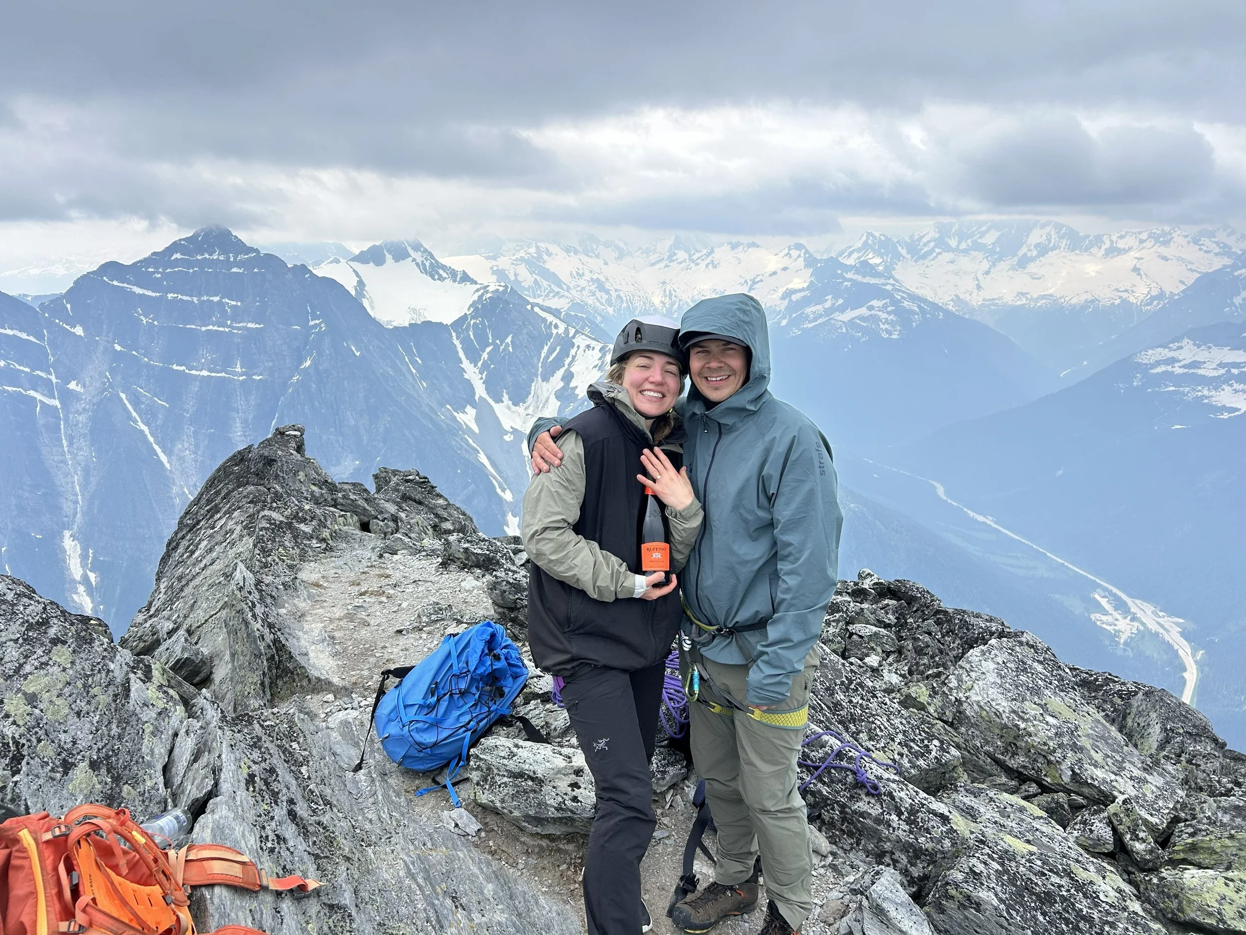 A smiling couple in outdoor gear standing on rocky mountain terrain, with snow-capped mountains and cloudy sky in the background.
