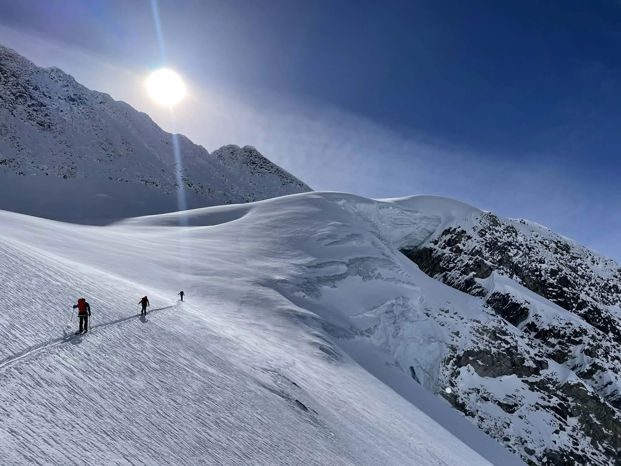 Snow-covered mountain landscape with a clear blue sky and bright sunlight. Three skiers are making tracks in the snow, heading up a slope.