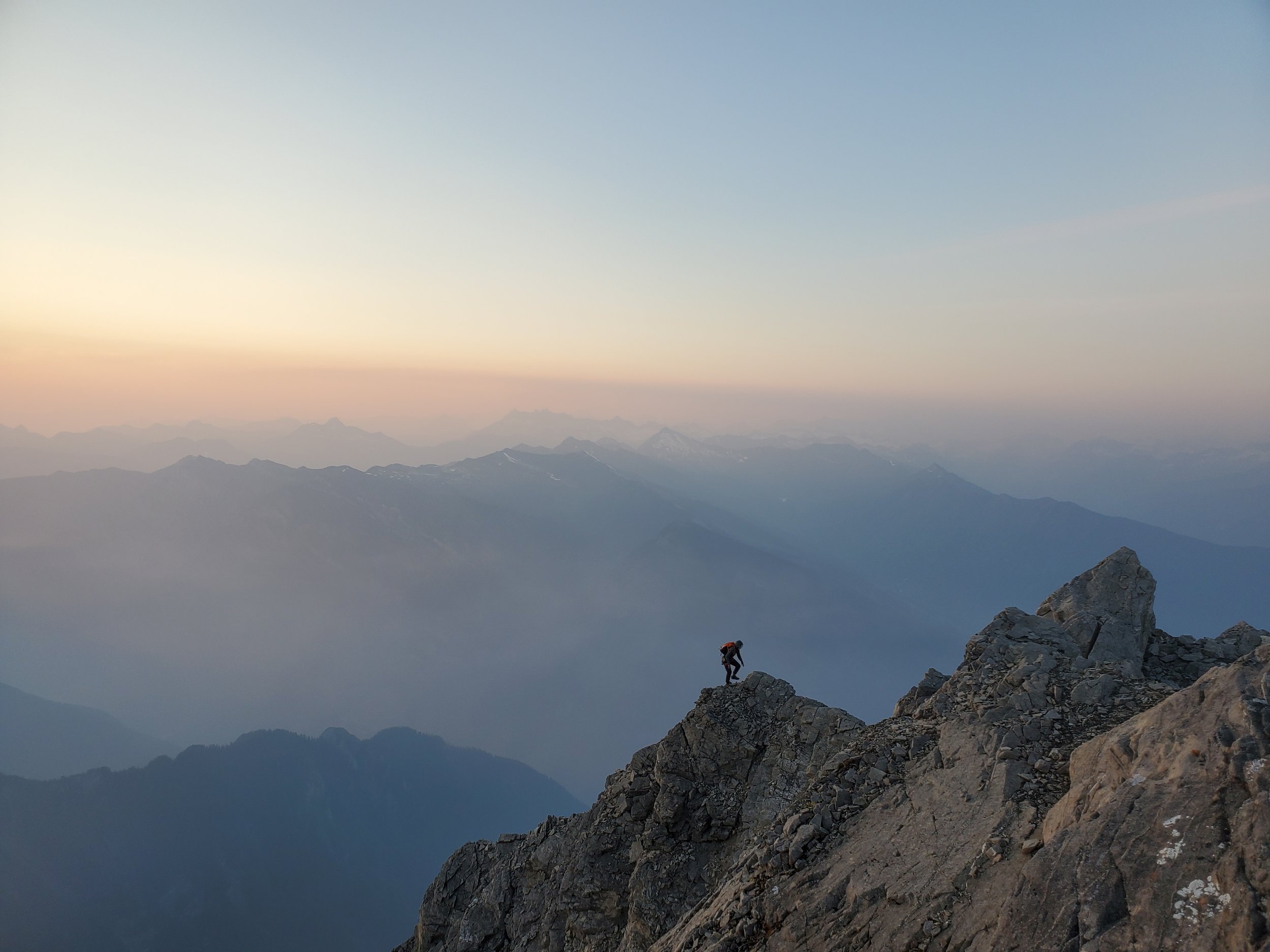 A person climbing a rocky mountain ridge during sunrise or sunset with distant mountain ranges in the background.
