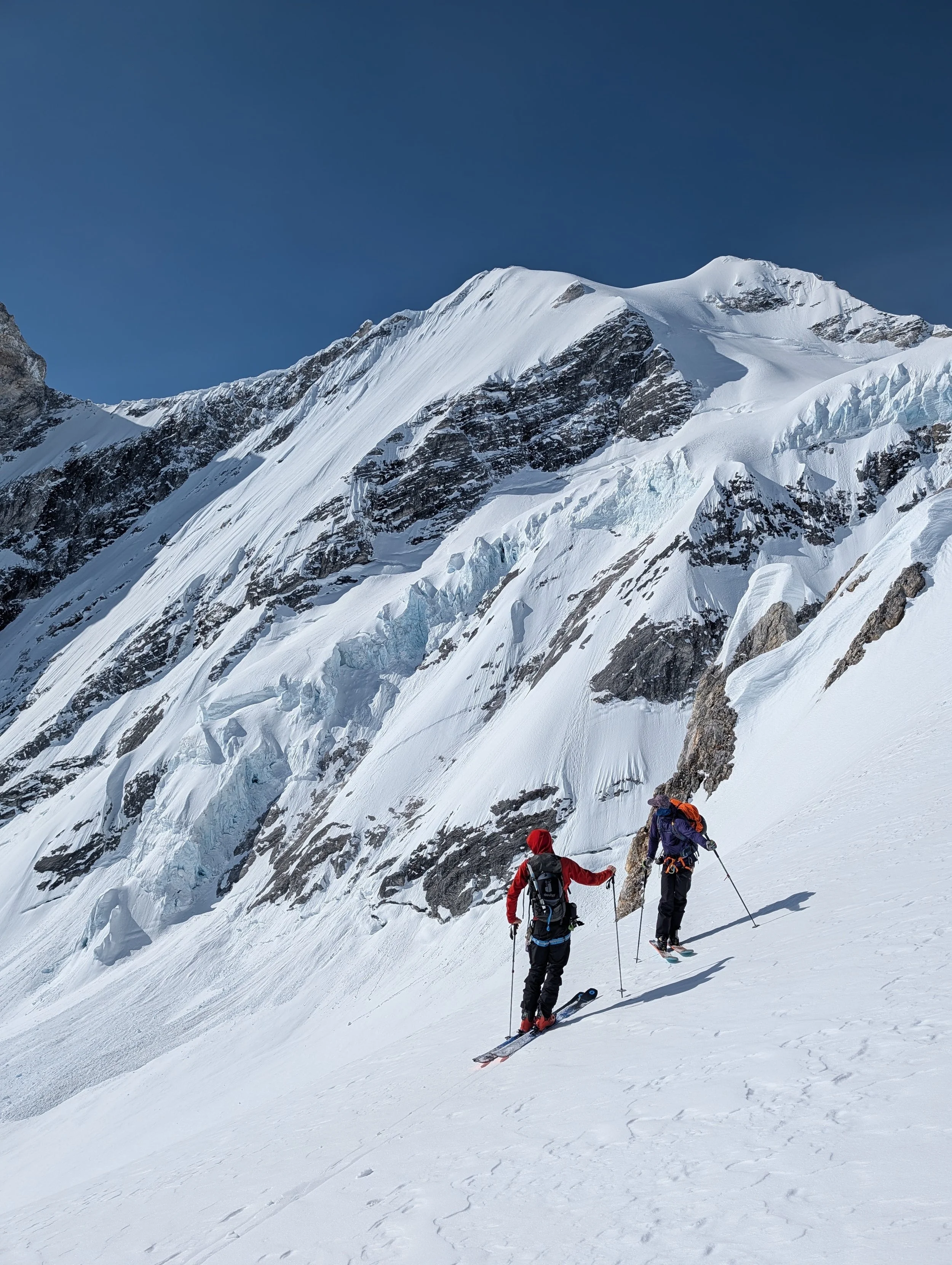 Two climbers ascending a snow-covered mountain slope with rugged peaks and glaciers in the background under a clear blue sky.