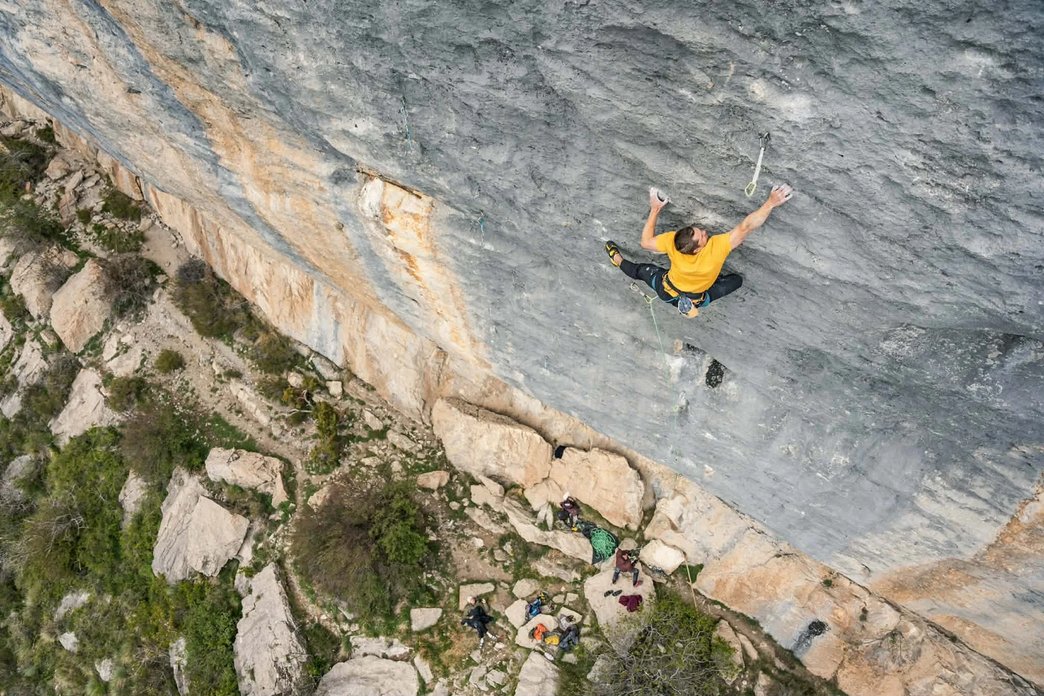 An aerial view of a person rock climbing a steep cliff, with equipment and other climbers below at the base of the cliff, surrounded by scattered rocks and greenery.