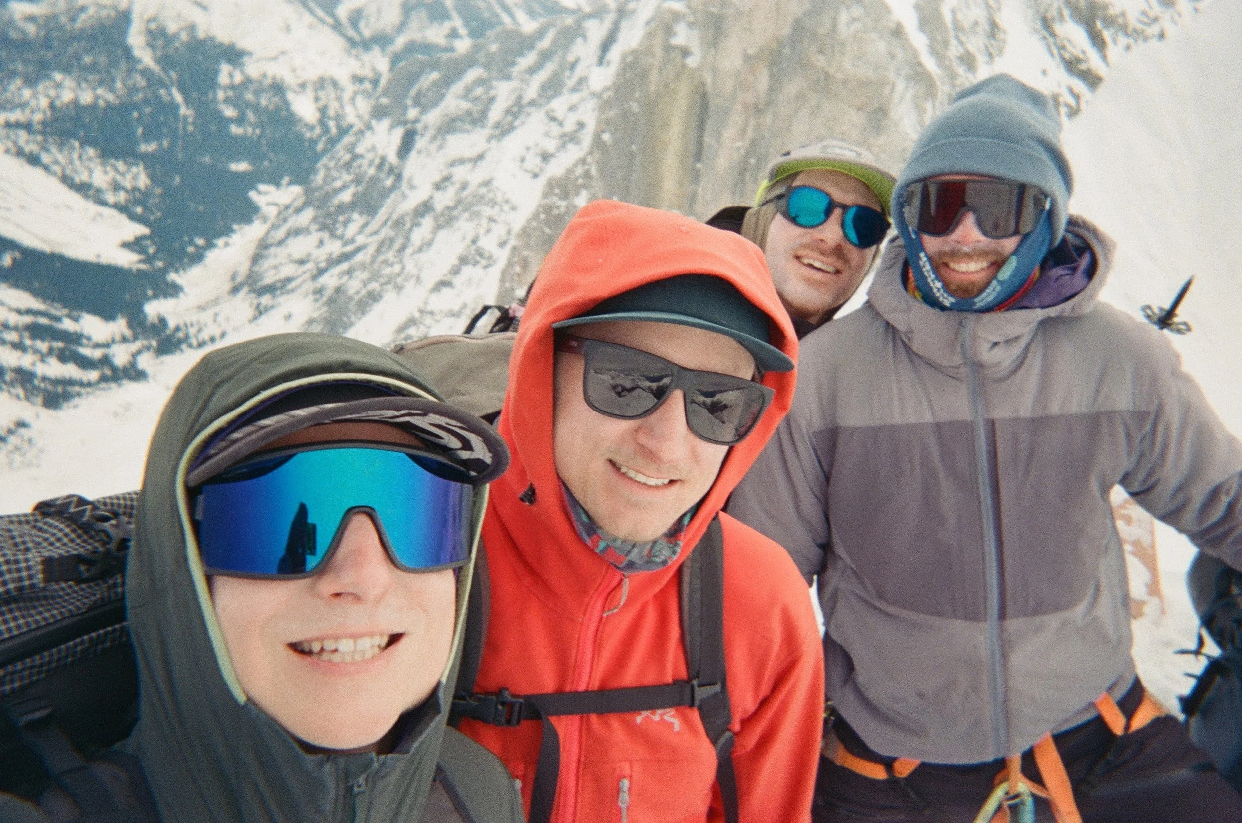 Four friends in snow-covered mountains wearing winter gear and sunglasses, taking a group selfie outdoors.