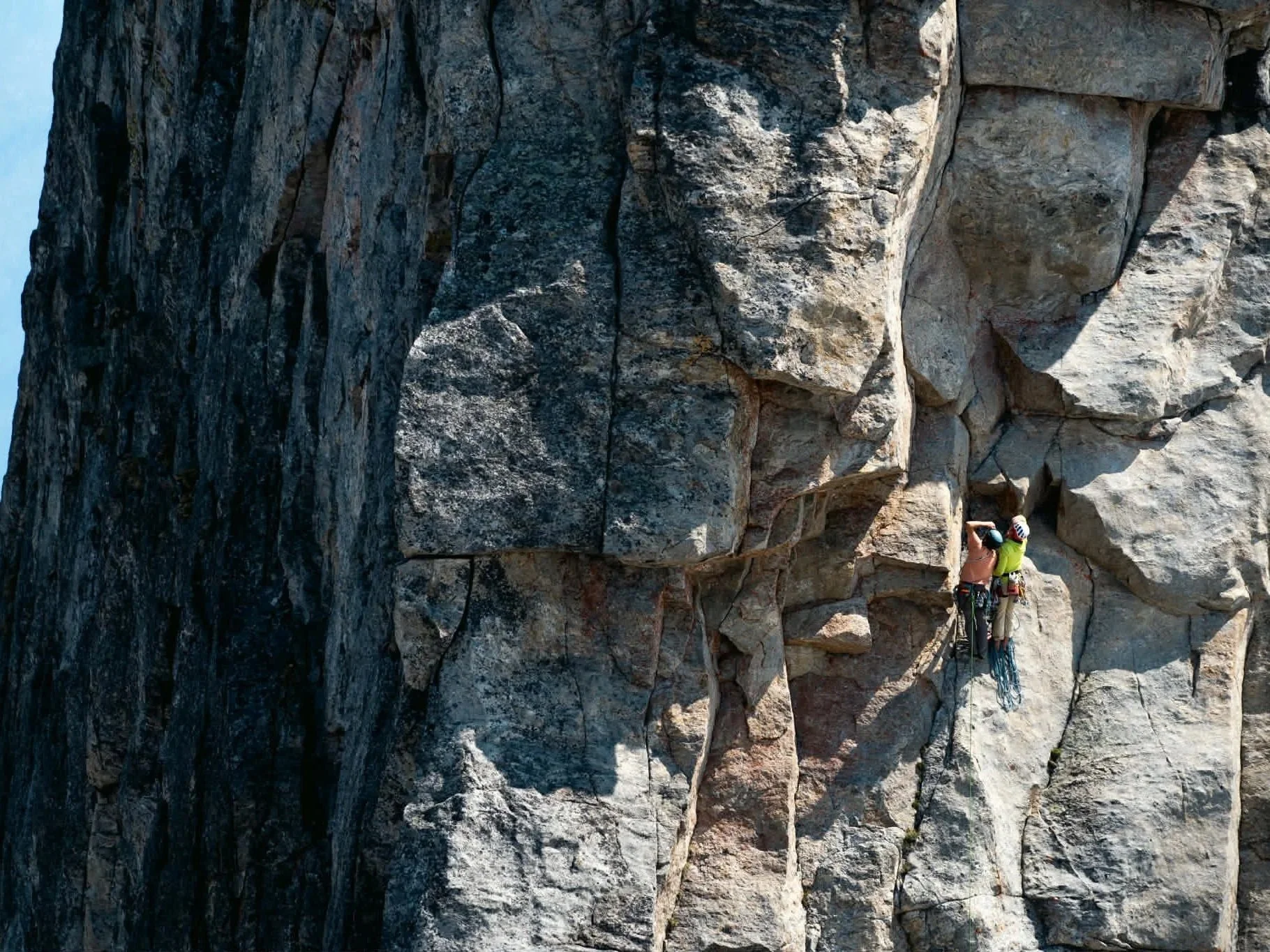 Two rock climbers with helmets and harnesses climbing a large steep rock face.
