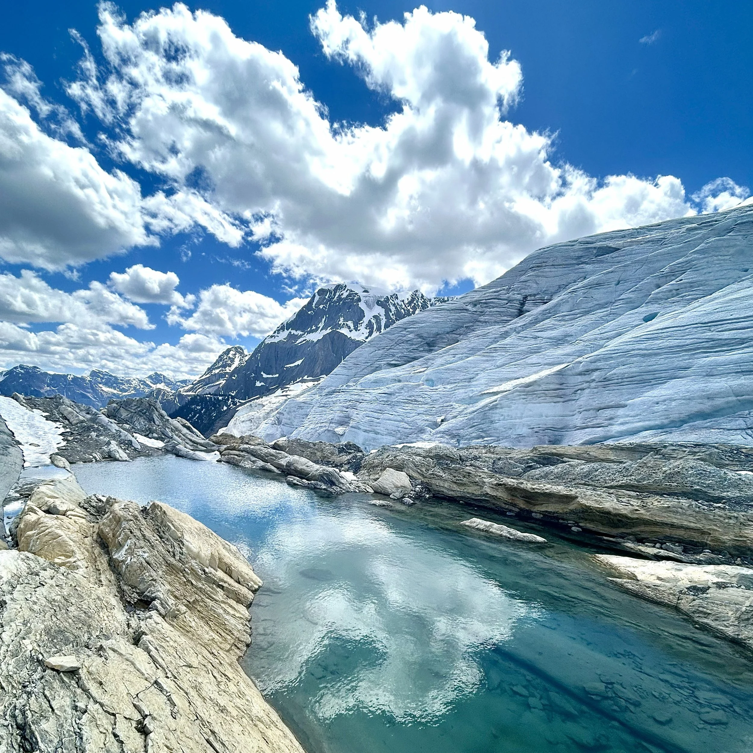 A glacier melting into a blue icy lake with rugged mountains in the background under a partly cloudy sky.