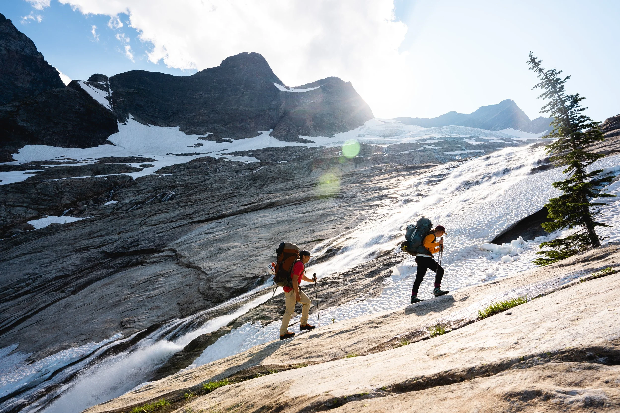 Three hikers with backpacks hiking through a snow-covered mountain landscape, with rocky terrain, a pine tree, and a bright sun in the sky.