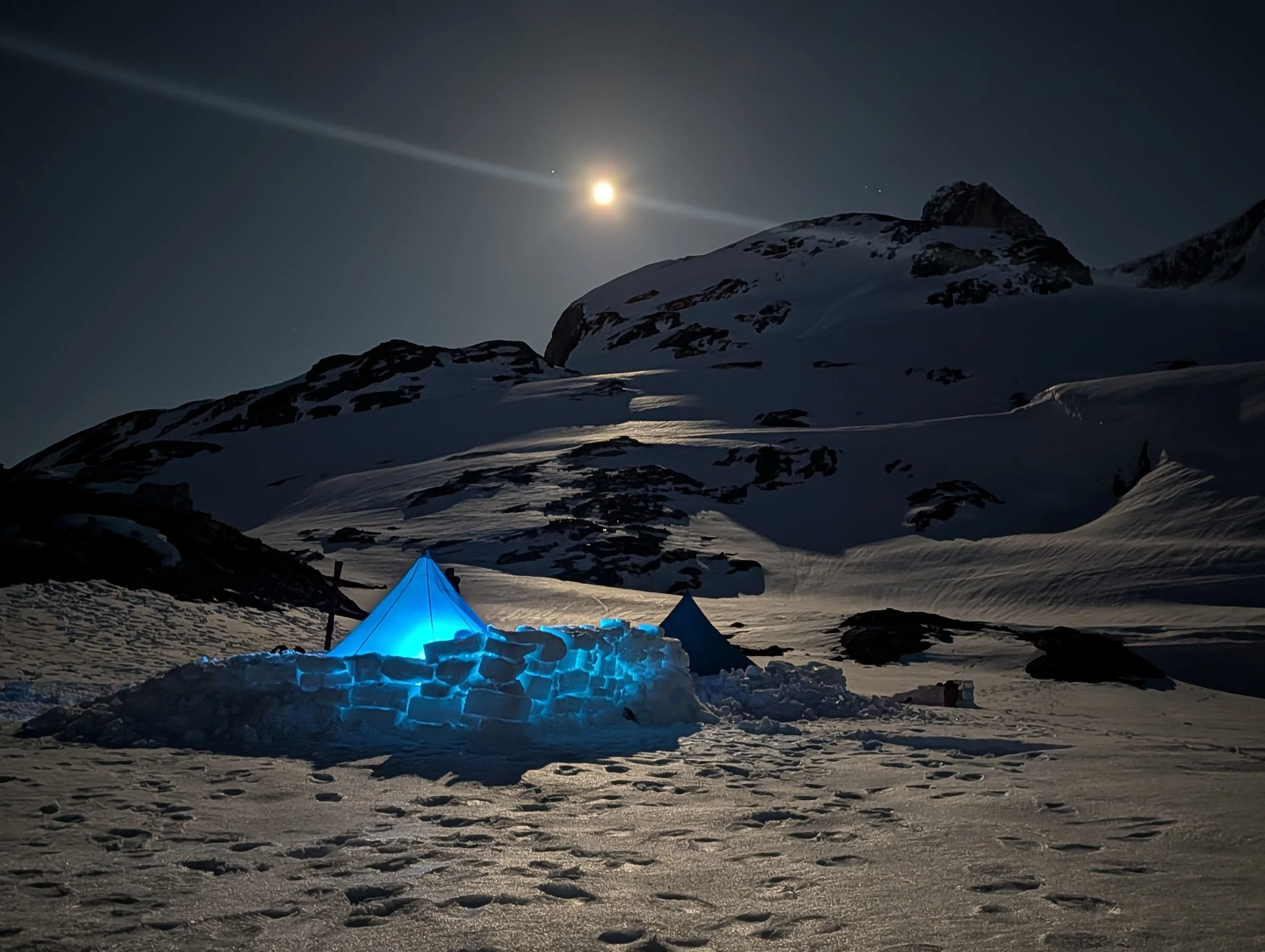 Nighttime scene of snow-covered mountains and a campsite illuminated with blue lights, with a full moon in the sky and stars visible.