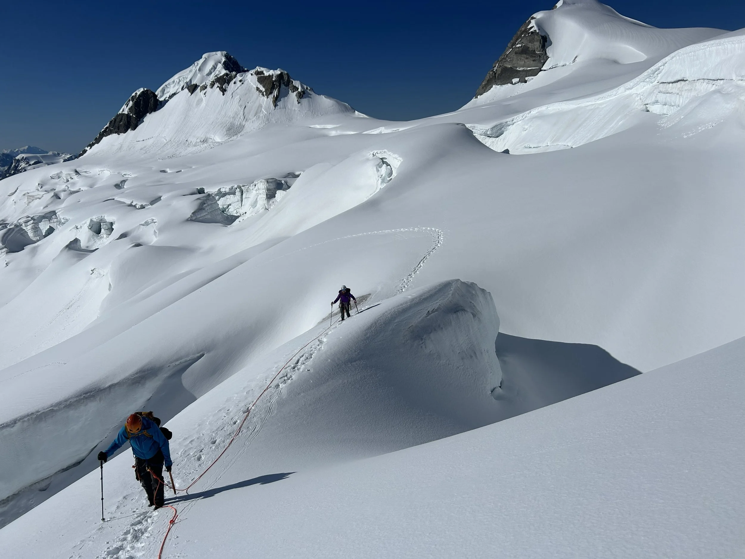Two climbers ascending a snow-covered mountain slope with towering snow and ice formations, under a clear blue sky.
