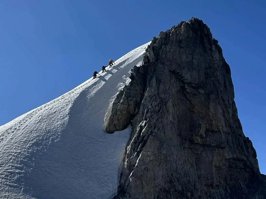 Three climbers ascending a snowy, steep mountain slope next to a large rocky cliff against a clear blue sky.