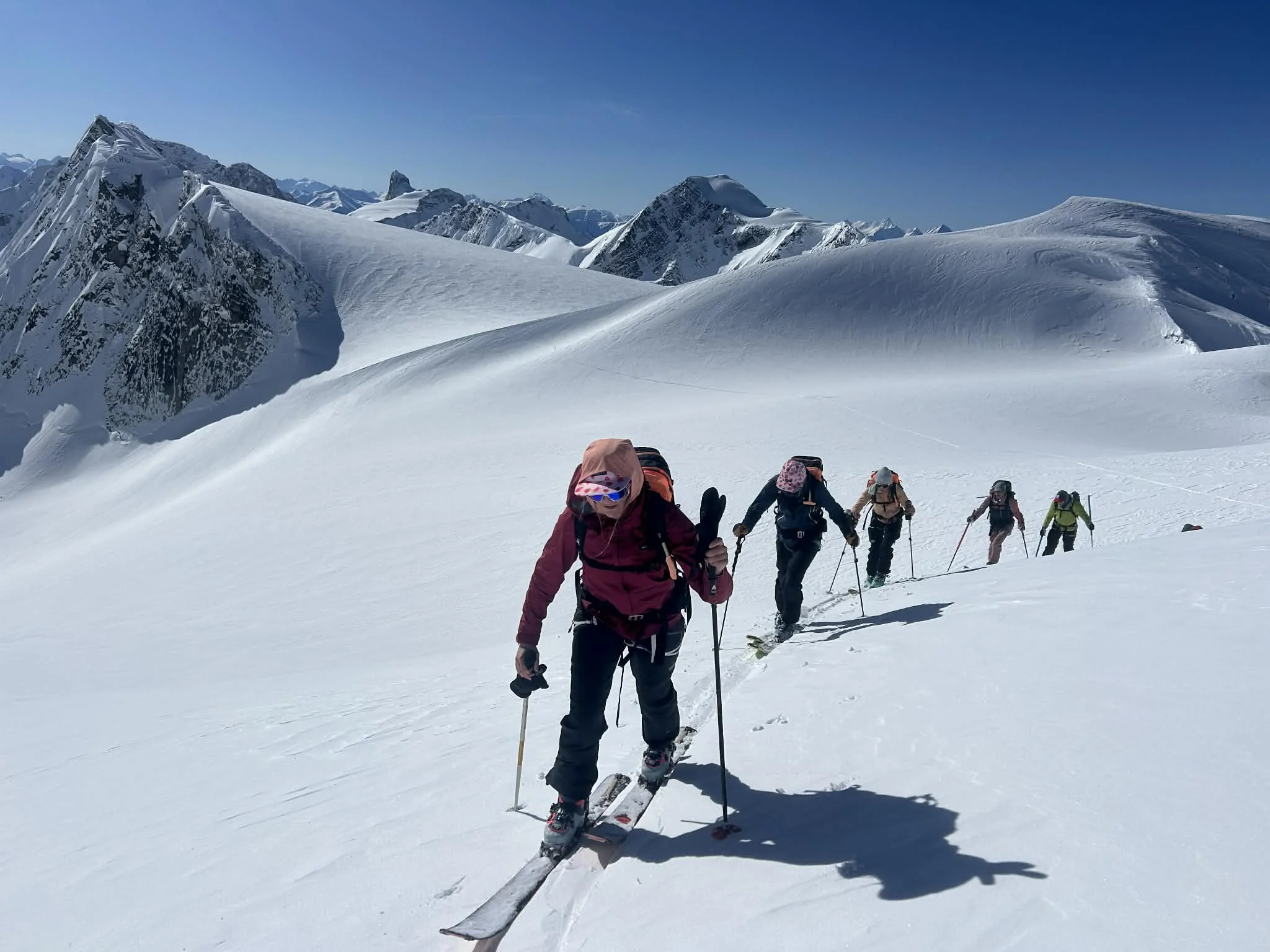 A group of skiers ascending a snowy mountain with snow-covered peaks in the background under a clear blue sky.
