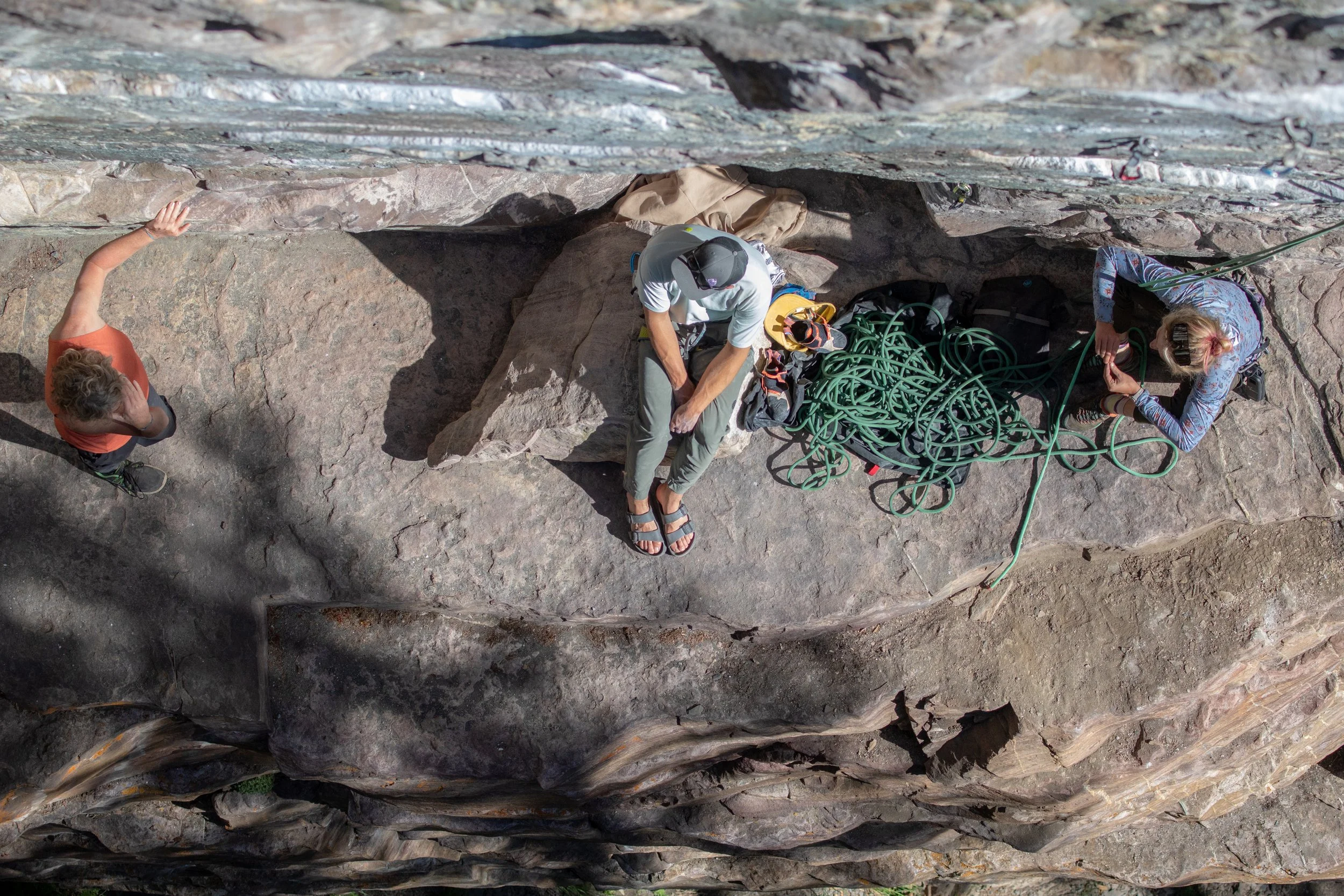 Two rock climbers preparing gear at the base of a steep rock face outdoors, with one resting on a ledge and the other adjusting climbing ropes.